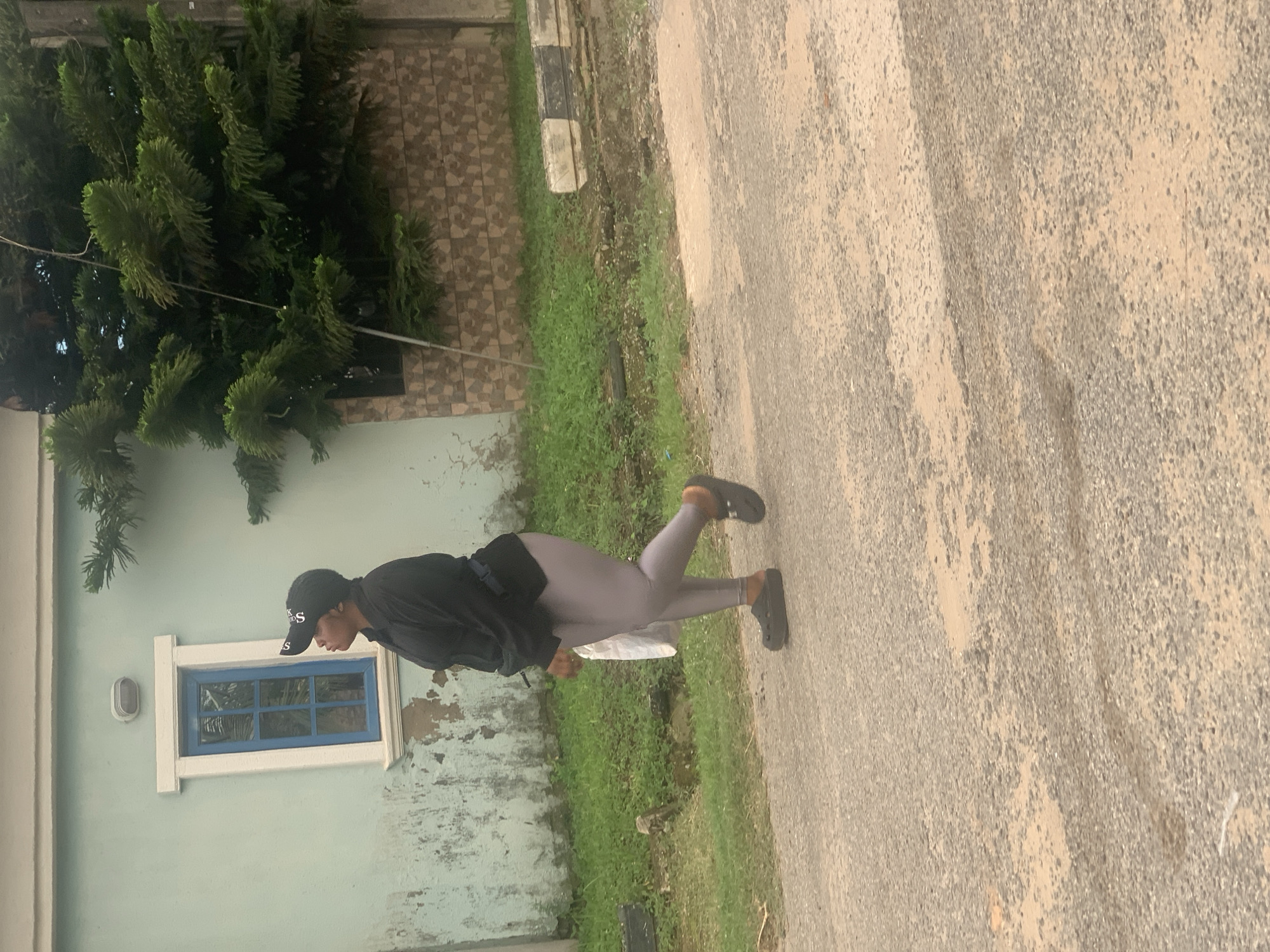 A full-body, side-profile shot shows a Black person wearing a black baseball cap, black long-sleeved shirt, and light grey leggings, walking on a paved road in Abuja, Nigeria. They are carrying a white plastic bag in their right hand and wearing black platform clogs. The person is walking from left to right, looking up and to the left. Behind them, a light blue building with a white-trimmed blue window and a light fixture is visible. To the right of the building is a dense evergreen tree and a textured stone wall. The ground is a rough, grey paved surface with some patches of sand and water. Green grass grows along the side of the road and a concrete curb with black and white markings is also present. The lighting suggests an overcast day.