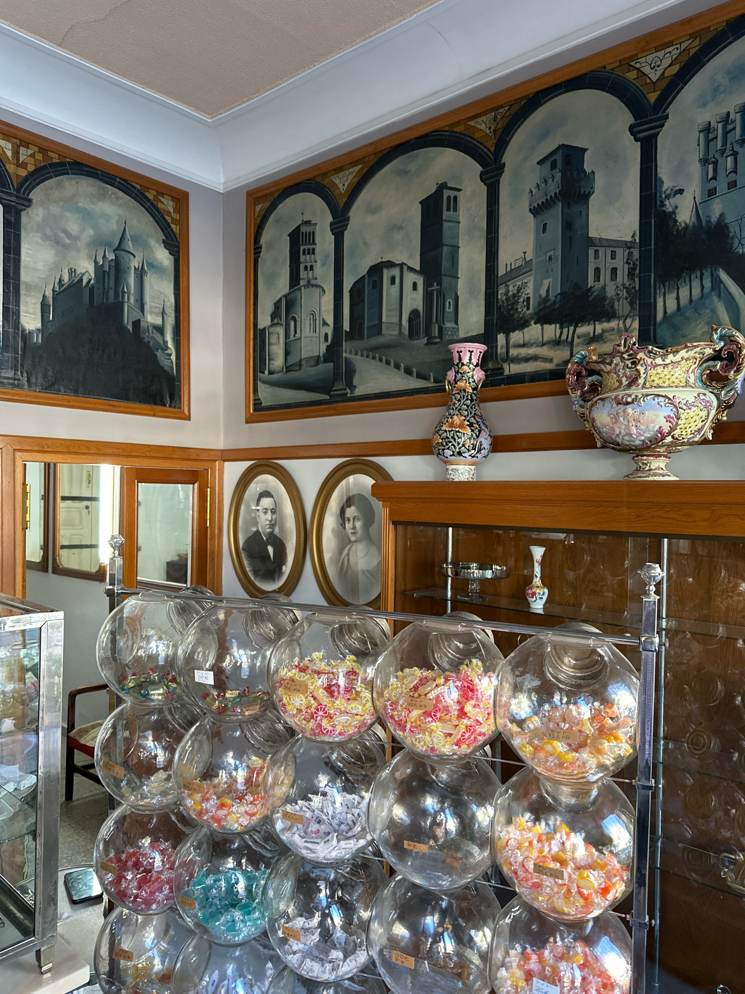 The image depicts the interior of a confectionery shop, likely a traditional establishment, with an emphasis on vintage decor and display. The walls are adorned with large arched murals depicting architectural scenes, possibly of castles or old buildings. In the foreground, a tiered display of clear glass globes filled with assorted wrapped candies is prominent. These globes are arranged in a staggered pattern, with the candies varying in color and type. Some globes have small price tags visible.
To the left of the candy display, a tall, narrow glass display case with a silver frame is partially visible, suggesting it holds other merchandise. In the background, a wooden cabinet or counter with glass-fronted shelves is present, displaying a few decorative items, including a small vase.
On the wall above the cabinet, two oval framed portraits of a man and a woman are displayed, adding to the nostalgic atmosphere. Between the portraits, the arched murals continue. A tall, decorative vase with a floral pattern sits on the top edge of the cabinet, to the right of the portraits. Next to it, a large, ornate ceramic vase with colorful painted figures and handles is a significant decorative element.
The overall lighting appears to be natural, coming from an unseen source, creating a bright and airy feel. The colors are muted, with a focus on the browns of the wood, the blues and grays of the murals, and the bright, varied colors of the candies. The scene evokes a sense of old-world charm and tradition.
While the image itself does not contain specific landmarks or clues to pinpoint the exact location within La Lastrilla, Spain, such a shop with its preserved aesthetic would contribute to the character of a historic district or a town that values its heritage.