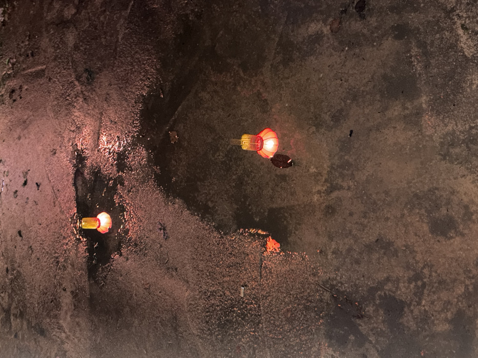 The scene depicts a dark, wet, and uneven paved surface in Ban Mai Saha Sampan, Thailand, at night. Two small, illuminated objects resembling floating lanterns are visible, each with a yellow upper portion and a red, rounded base emitting a warm reddish-orange light. One object is positioned in the upper-left quadrant of the frame, partially submerged in a puddle, its light reflecting on the water. The second, larger object is located in the lower-right quadrant, also within a reflective puddle, casting a similar glow. The ground exhibits various wet patches and puddles, indicating recent precipitation or standing water, reflecting the ambient darkness and the red light sources. Scattered debris, possibly organic matter like leaves, is present on the surface and within the puddles. No individuals are visible. The overall lighting is dim, relying primarily on the internal illumination of the two floating objects.