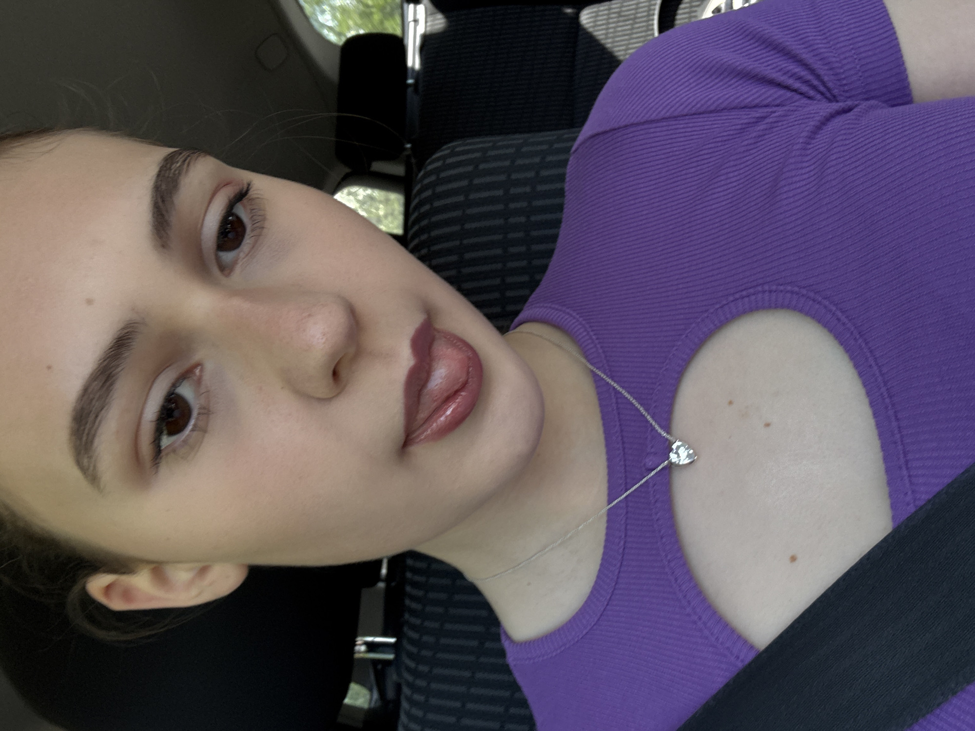 A close-up, tilted selfie of a young woman in a car, presumably in Levski, Bulgaria. She has fair skin, brown eyes, and arched eyebrows. She is wearing a purple ribbed top with a cutout near the collarbone and a silver necklace with a heart-shaped pendant. A black seatbelt is visible across her chest. The car seat is upholstered in a dark gray patterned fabric. Through the car window, a glimpse of greenery and trees can be seen. Her lips are glossy, and her tongue is slightly visible.