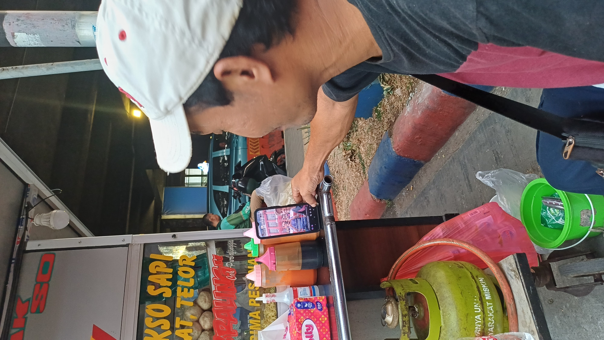 A man wearing a white baseball cap with red accents is holding a smartphone in his right hand, viewing a video on the screen. He is positioned in the foreground, on the right side of the frame, and is looking towards the phone. His left arm is partially visible, and he is wearing a dark t-shirt with a maroon stripe across the chest.
Behind him, to the left, is a food cart. The cart has a white structure with red and yellow lettering that appears to be in Indonesian. Visible text includes "AKSO SAPI" and "AT TELOR". Several bottles of sauces and a box of tissues are on the cart's surface. A green propane tank is also visible on the lower left of the cart.
In the background, the scene appears to be an outdoor urban environment. There is a road with a teal-colored vehicle and a motorcycle. A person in a green shirt is seen near the teal vehicle. The area also has a concrete barrier painted with red and blue stripes, and some foliage. The lighting suggests it might be evening or dusk. The specific location in South Jakarta, Indonesia is not explicitly identifiable from the image itself, but the presence of Indonesian language on the cart and general urban Southeast Asian aesthetics are evident.