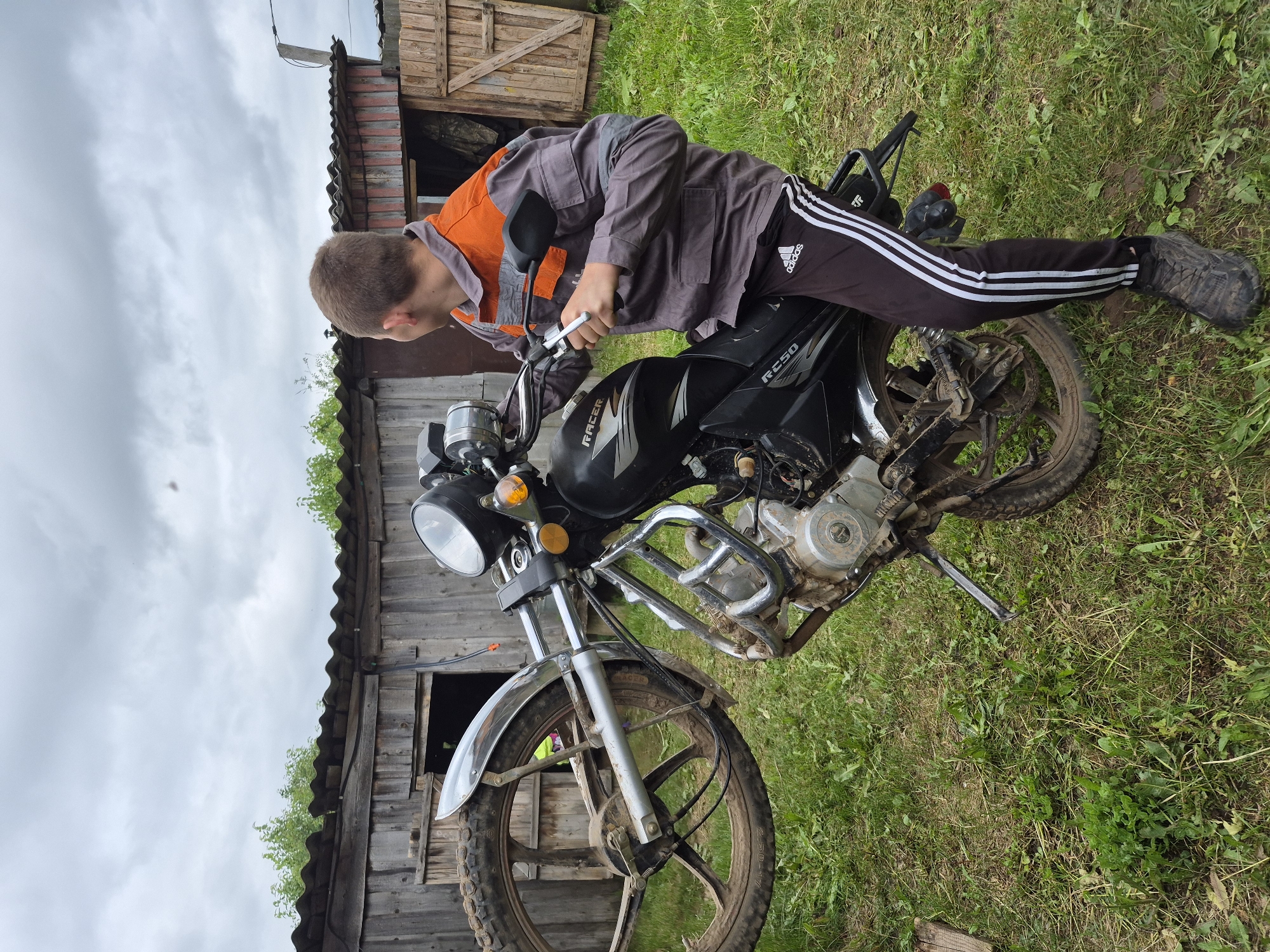 In Kharnavy, Russia, an outdoor scene captures a young male astride a black "RACER RC250" motorcycle on a cloudy day. The individual, seen from behind and looking towards the right, is lifting the motorcycle's front wheel high off the ground, holding the handlebars as if attempting a wheelie. He is dressed in a grey long-sleeved work jacket with orange shoulders, black Adidas track pants featuring white stripes, and dark, mud-splashed athletic shoes. The motorcycle's front wheel, suspension, and lower engine components are heavily covered in mud. In the background, a weathered wooden shed with a corrugated roof is visible, surrounded by sparse green trees and bushes, while the ground consists of grass mixed with dirt and weeds. The sky is overcast and grey.
