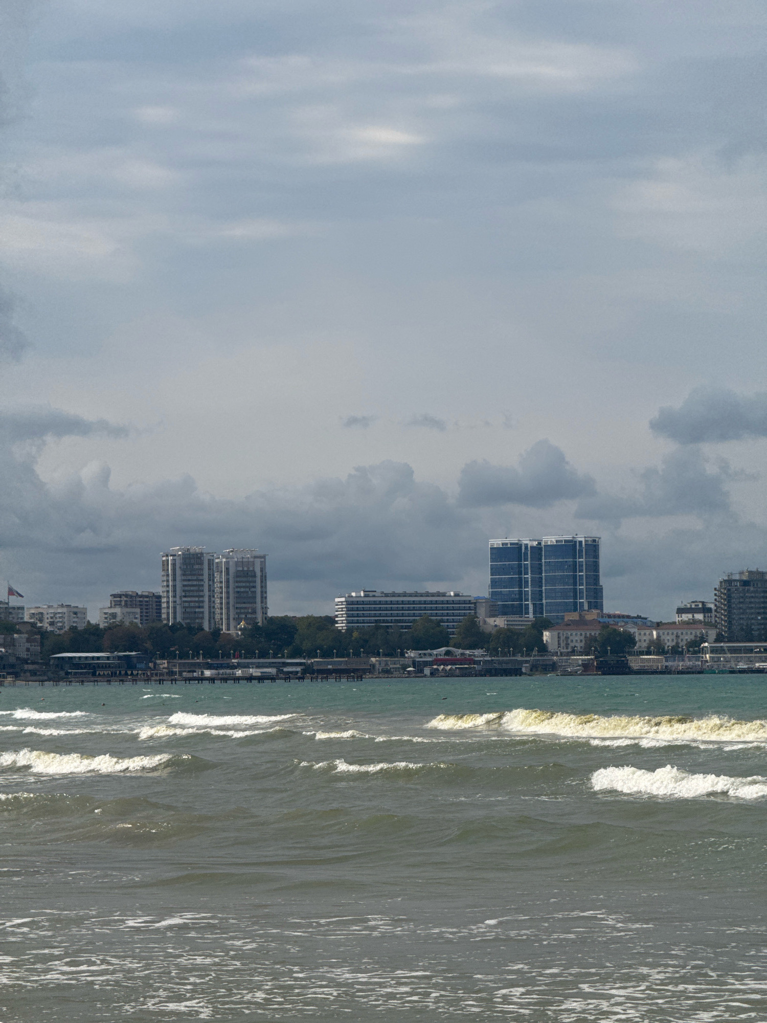 The image depicts a cloudy day at the Black Sea coast in Anapa, Russia. In the foreground, gentle waves are rolling onto a beach, with white foam visible along the shore. In the mid-ground, a long pier stretches out over the water, lined with various buildings and structures, suggesting a developed waterfront area. Behind the pier, a cityscape unfolds with several tall buildings, including modern high-rises with glass facades and older, more traditional buildings. Lush green trees separate the waterfront from the city, and the sky is filled with heavy, grey clouds, creating an overcast atmosphere. The overall scene conveys a sense of a coastal city on a somber day.