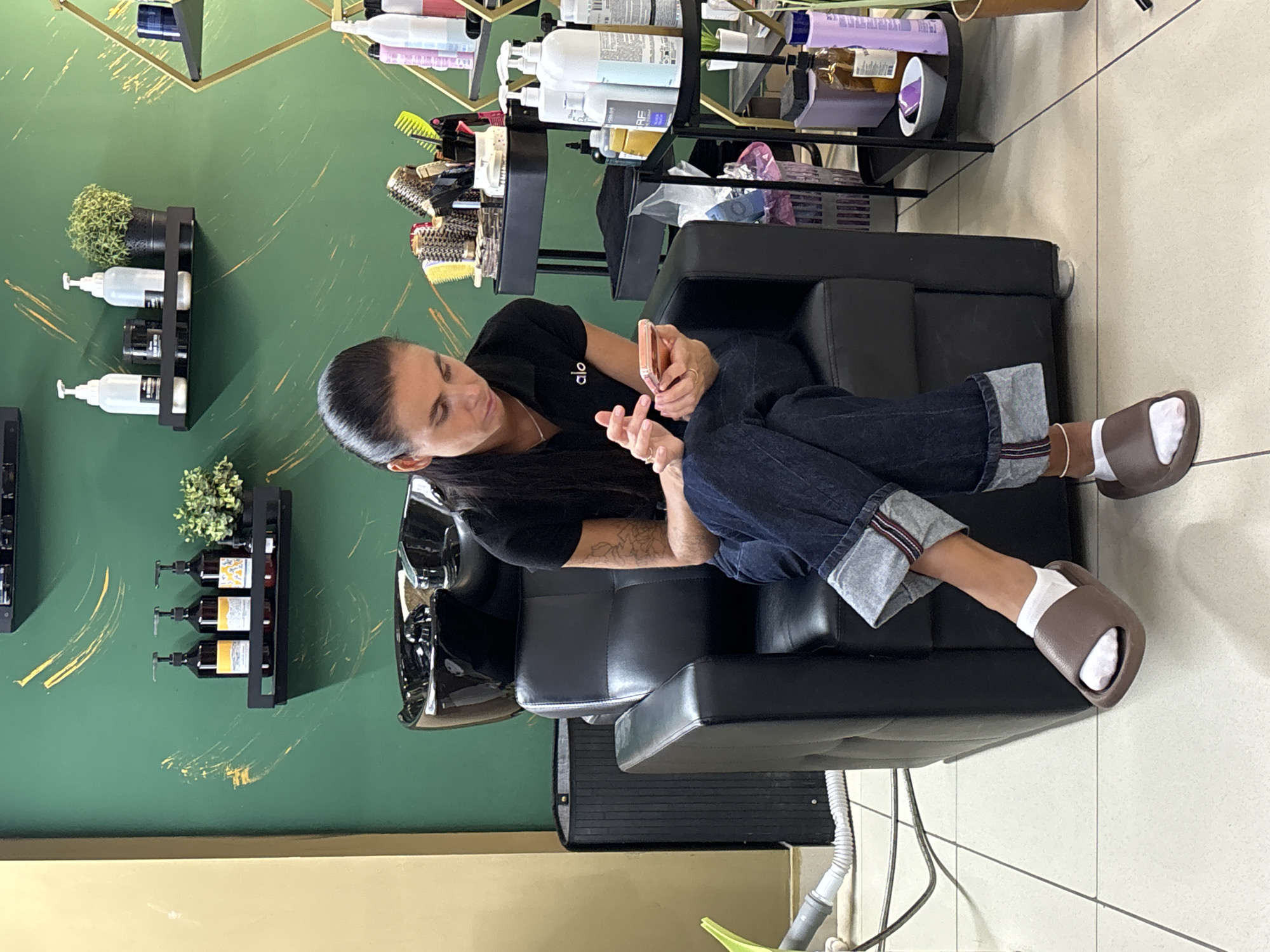 A woman is sitting in a salon chair, looking at her phone. She is wearing a black shirt, blue jeans, white socks, and brown sandals. The salon has green walls with gold accents, and shelves with hair products and plants. The location is in the city of Chaikovsky, Russia.