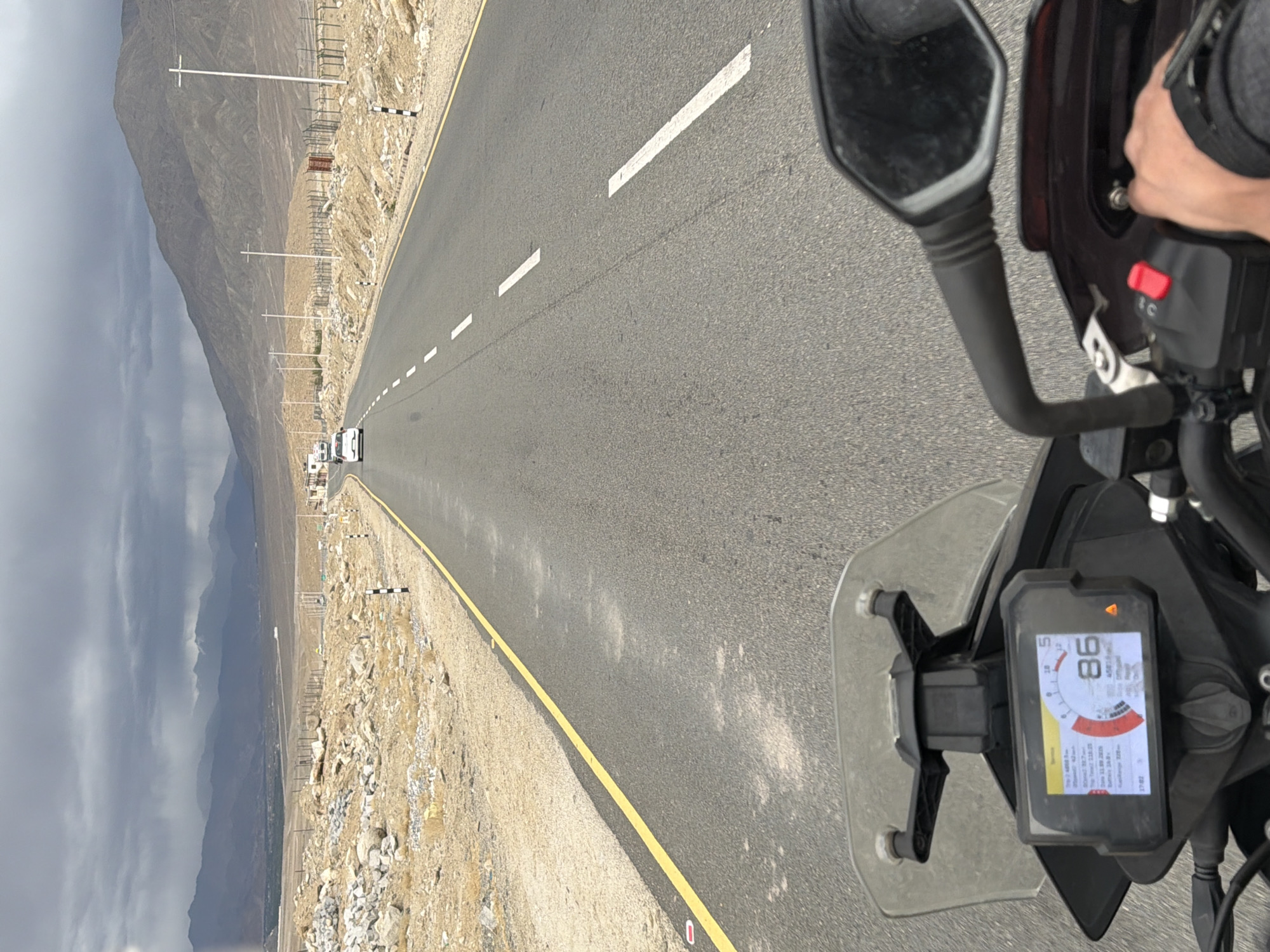 The image is a point-of-view shot from a motorcycle traveling on a paved road through a barren, mountainous landscape. The road has a yellow line on the left and white dashed lines indicating lanes. In the distance, there are mountains under a cloudy sky. There are a few white vehicles on the road ahead. The motorcycle's dashboard is visible in the foreground, showing a speed of 86. The environment appears to be a remote area in India, possibly near Chumkar, characterized by rocky terrain and sparse vegetation. A hand is visible on the handlebar of the motorcycle.