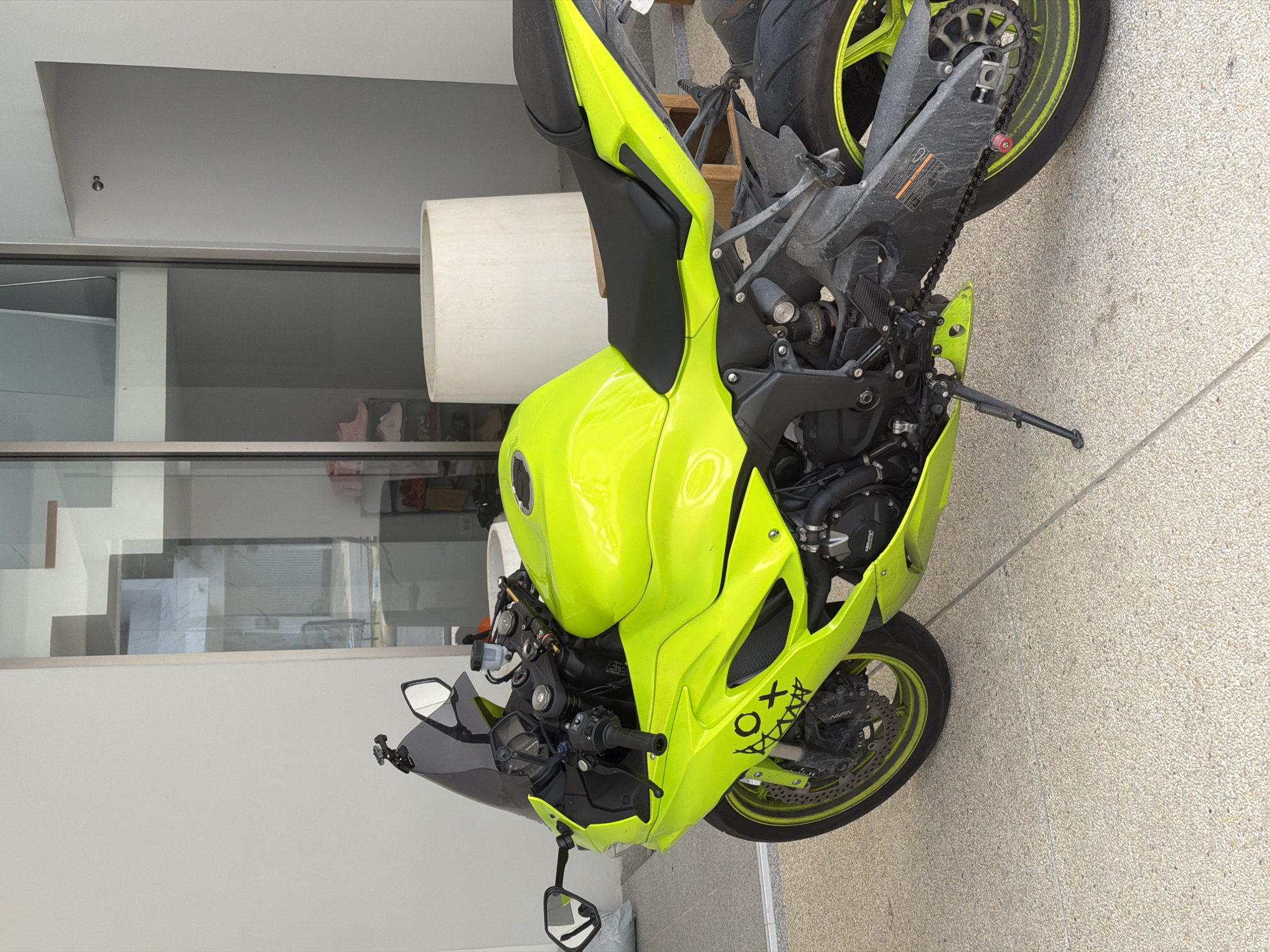 An electric yellow sport motorcycle is parked on a light-colored, speckled concrete surface. The motorcycle is positioned at an angle, facing towards the right of the frame. The front wheel, handlebars, mirrors, and a portion of the fairing are visible on the left. A dark tinted windscreen is mounted on the front. The body of the motorcycle is a bright, fluorescent yellow, with black accents on the seat, handlebars, and some mechanical components. A black sticker with a stylized "OX" and jagged lines is present on the lower front fairing. The rear section of the motorcycle, including the seat, rear wheel with a yellow rim accent, and swingarm, is visible on the right. The background consists of a modern building with large glass doors and windows, reflecting the surroundings. To the right of the motorcycle, a tall, cylindrical white object, possibly a planter or a decorative element, is partially visible. The scene appears to be an outdoor area adjacent to a building, potentially a residential or commercial property. No people are present in the image.