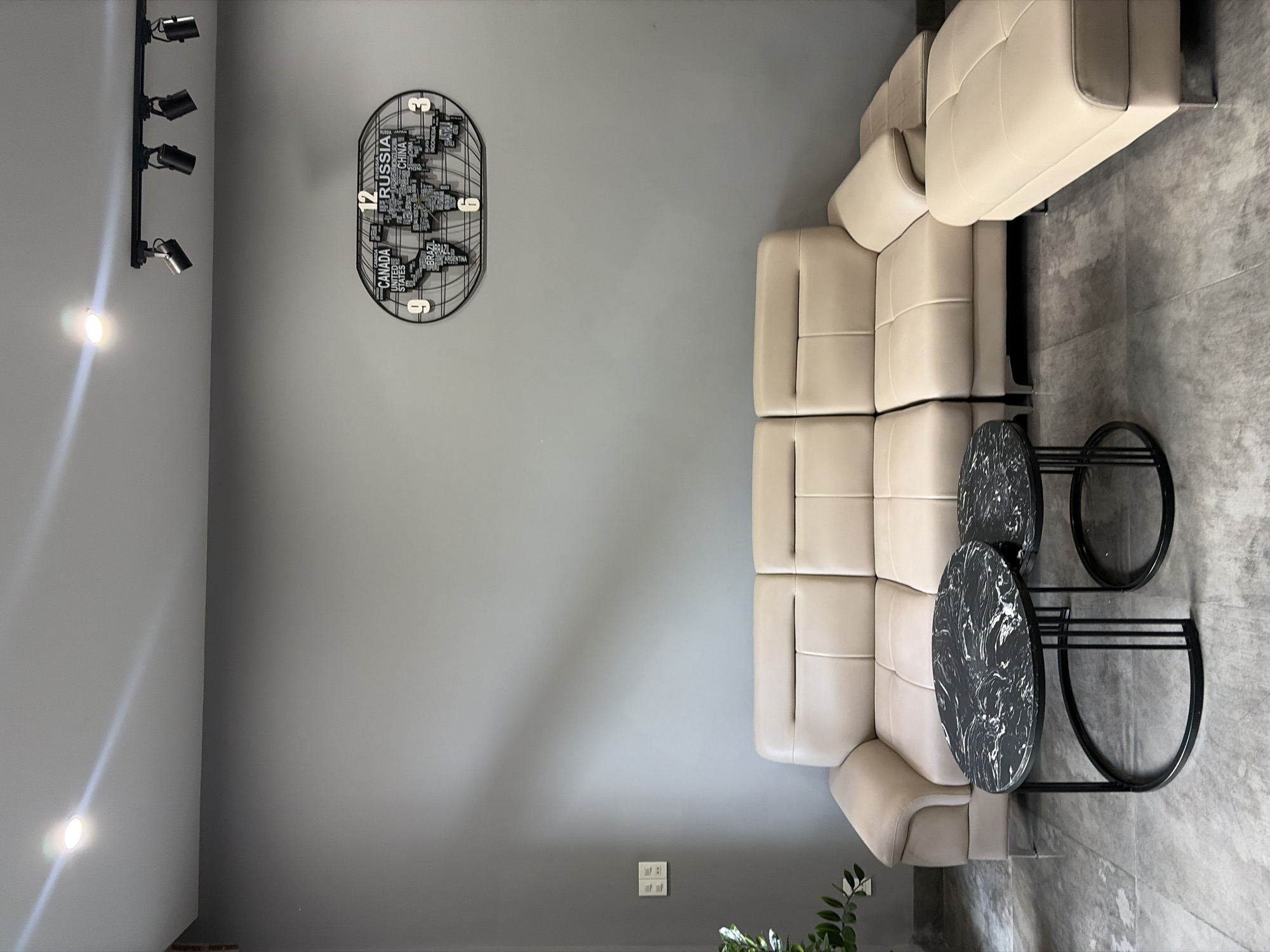 The image depicts a modern living room interior, likely situated in a residence or showroom in Da Nang, Vietnam. The room features a light beige sectional sofa with multiple seating sections and a matching ottoman. In front of the sofa, two round nesting coffee tables with black marble tops and black metal legs are placed. The walls are painted a neutral gray, and the floor is tiled with large, gray concrete-look tiles. Above the sofa, a decorative black metal clock shaped like a world map is mounted on the wall. Track lighting is visible on the ceiling, with several adjustable spotlights directed downwards. A small section of a green plant is visible in the bottom left corner, and a light switch plate is mounted on the wall to the left of the sofa. The overall ambiance is contemporary and minimalist, with the gray and beige tones creating a sophisticated and clean aesthetic. There are no people present in the scene.