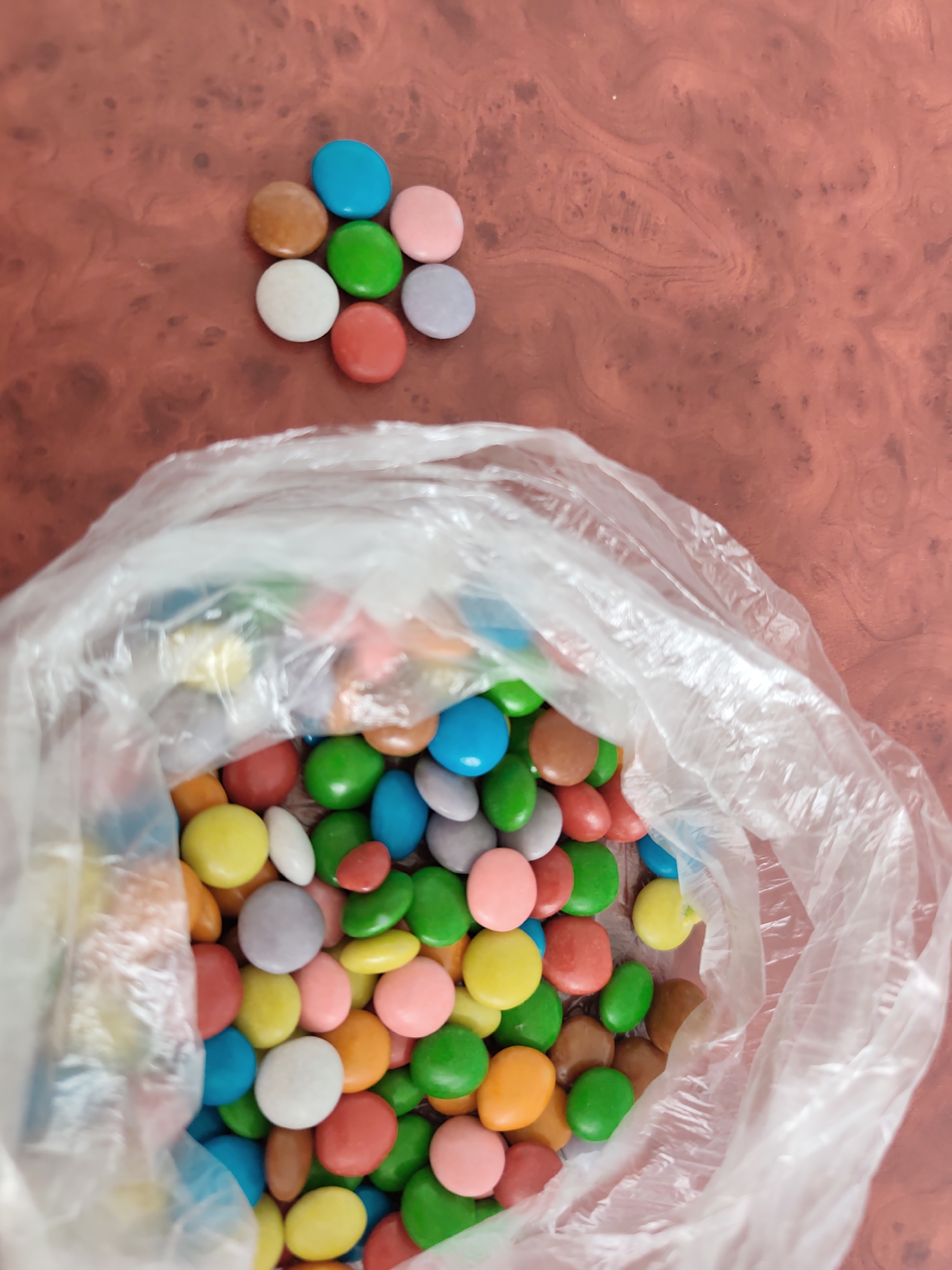 A high-angle, overhead photograph captures a collection of multi-colored, disc-shaped candies on a reddish-brown, marbled surface. The scene is situated in the city of Chaikovsky, Russia.
In the upper left-center of the frame, seven individual candies are arranged in a loose cluster, exhibiting distinct colors: brown, light blue, green, pink, white, red, and light purple. The majority of the image is dominated by a clear, crinkled plastic bag, positioned in the lower half to two-thirds, from which a dense assortment of similar multi-colored candies spills forth. These candies display a wide spectrum of hues including red, green, blue, yellow, orange, pink, white, brown, and light purple. The plastic bag appears translucent and somewhat crumpled.
The background surface is a consistent reddish-brown with intricate, darker, swirling patterns that suggest a wood grain, polished stone, or resin finish. The lighting is even, casting minimal shadows. No people, discernible actions beyond the presence of the objects, or specific interactions are visible within the frame.
