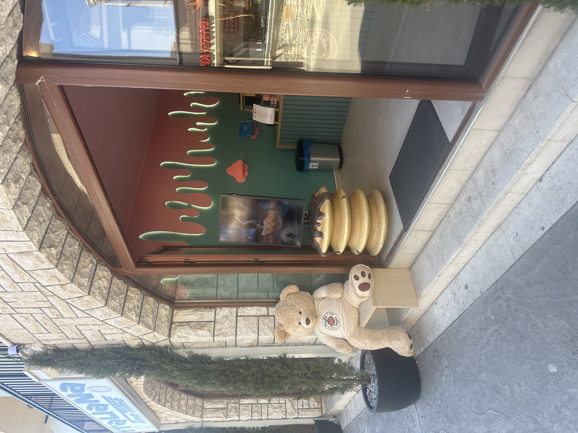 The image shows the entrance to "La Parisienne" bakery in Budva, Montenegro. The entrance features a brown-framed glass door. Inside, the walls are decorated with a pink upper section and a green lower section with stylized dripping shapes and clouds. A TV screen displays moving images. A row of golden-painted, doughnut-shaped objects are positioned next to the entrance. Outside, a large teddy bear wearing a branded t-shirt is seated on a small wooden bench next to a potted evergreen plant. The exterior is tiled with stone bricks and a blue and white sign for the bakery is visible. The ground is paved with concrete tiles.