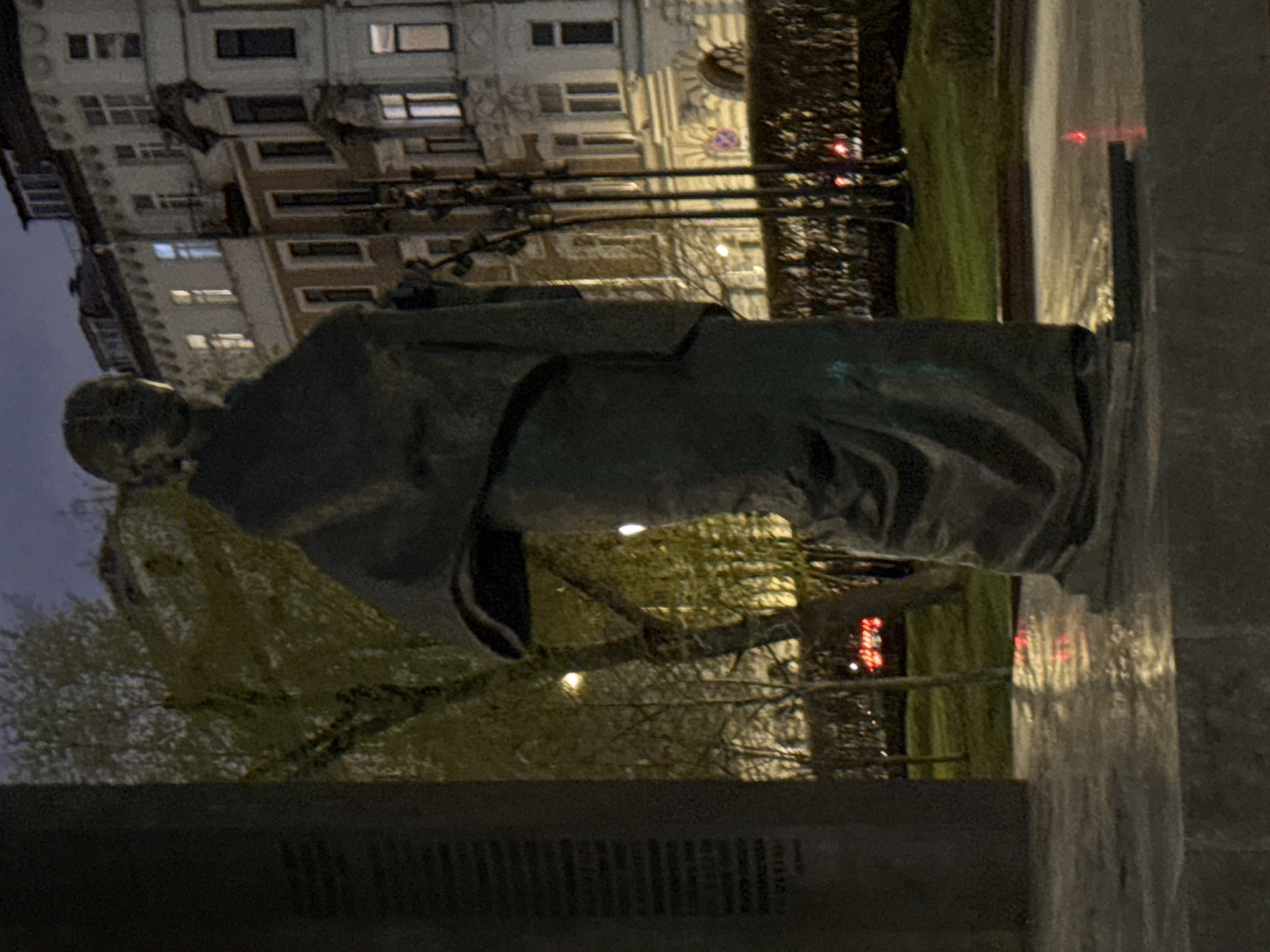 The image depicts a bronze statue of a seated man in Moscow, Russia. The statue is located on a stone plinth. Behind the statue is a building with multiple windows and street lamps. There is a tree to the left of the statue. The lighting suggests it is evening or night.