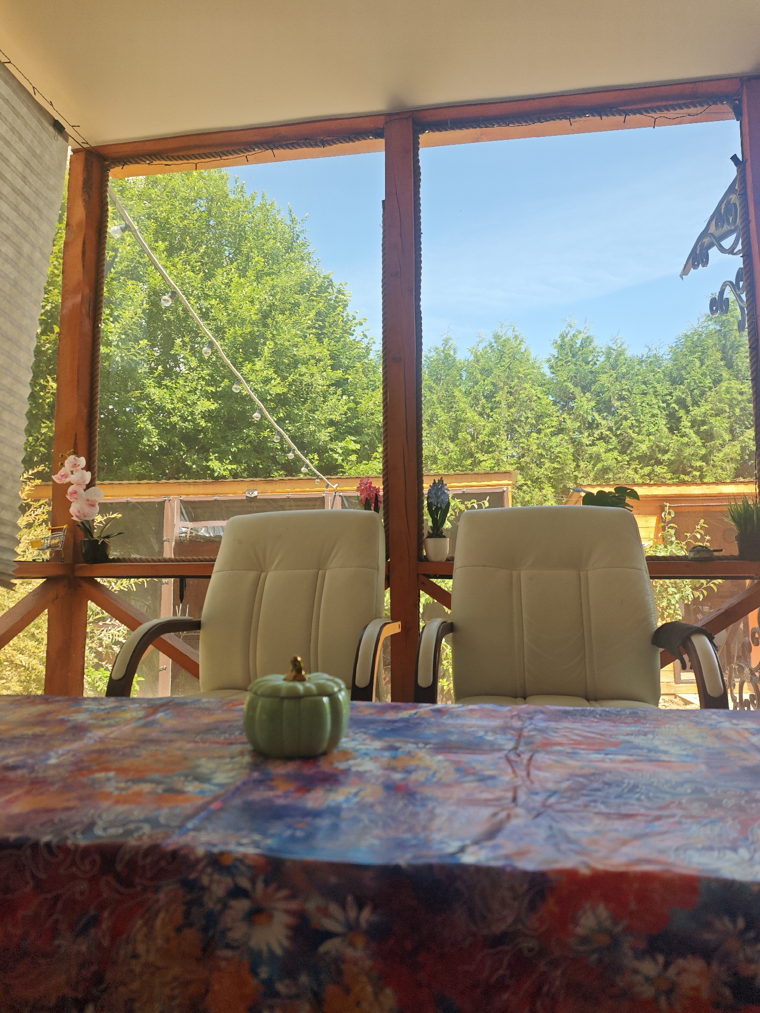 The image depicts a screened-in porch with two upholstered chairs facing a table covered with a floral-patterned tablecloth. A small green pumpkin-shaped decoration sits on the table. Through the screen, a lush green forest is visible under a clear blue sky, suggesting a rural or suburban setting. The porch has wooden posts and railings, and string lights are visible along the ceiling. The overall impression is one of a tranquil outdoor space. The specific location of Хлевино, Russia, is not discernible from the visual content of the image.