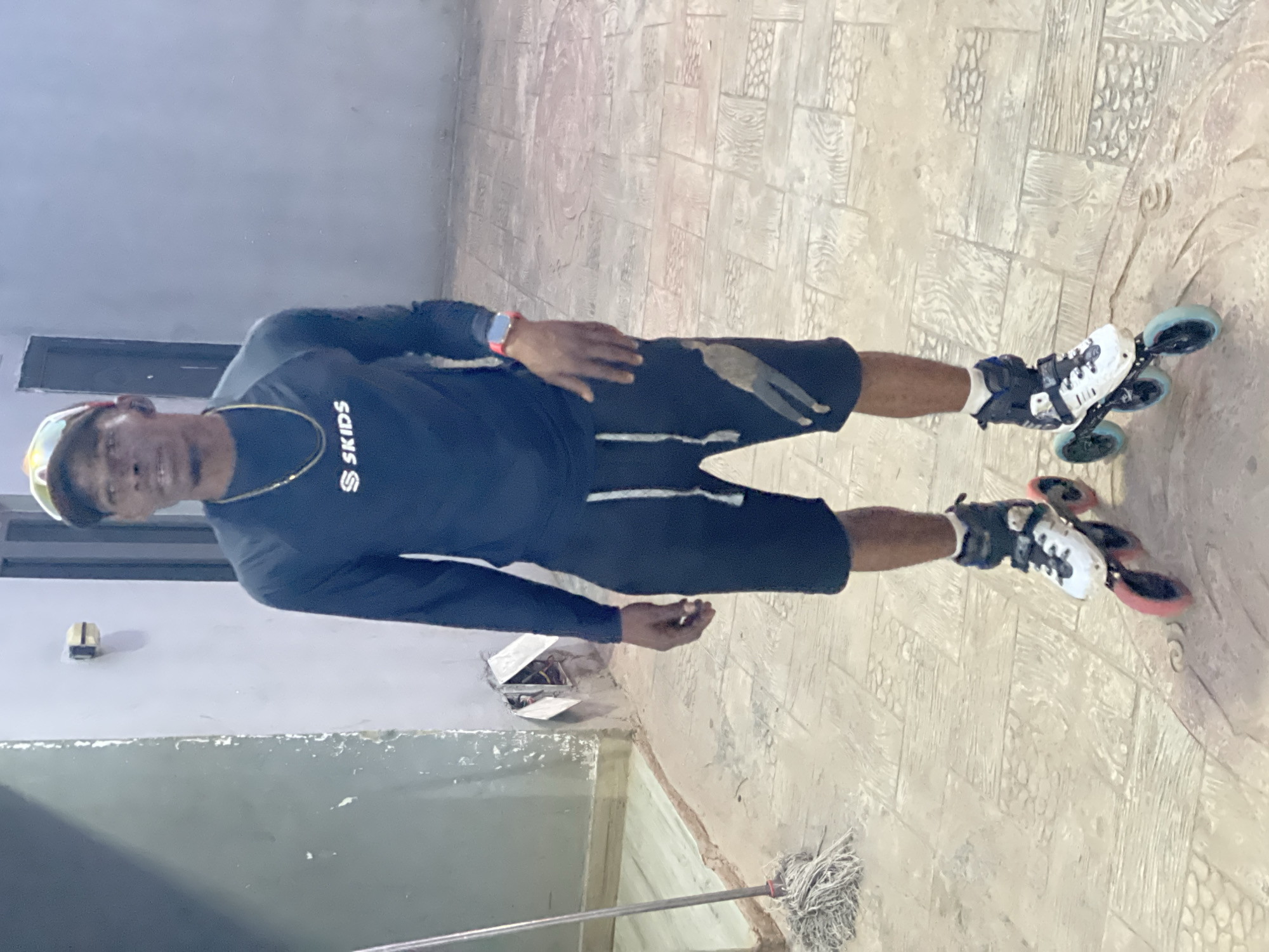 A full-body portrait captures a male individual standing on a patterned floor, looking directly at the camera with a slight smile. The scene is set in what appears to be a brightly lit outdoor or covered area in Akwanga, Nigeria. The man has dark skin and short dark hair, and wears a black long-sleeved shirt with the white logo "S SKIDS" on the chest, along with black shorts that have a white stripe. A cap with sunglasses resting on it is on his head, and a gold chain adorns his neck. He is wearing inline roller skates; one has blue/teal wheels, and the other has pink/red wheels. A red smartwatch is visible on his left wrist. The ground is a light-colored, possibly stamped concrete or tiled surface featuring various textures, including patterns resembling wood grain and pebbles. In the background, there is a plain light-colored wall with a darker rectangular opening or architectural feature to the left. A string mop with a metal handle is partially visible on the floor near his feet.