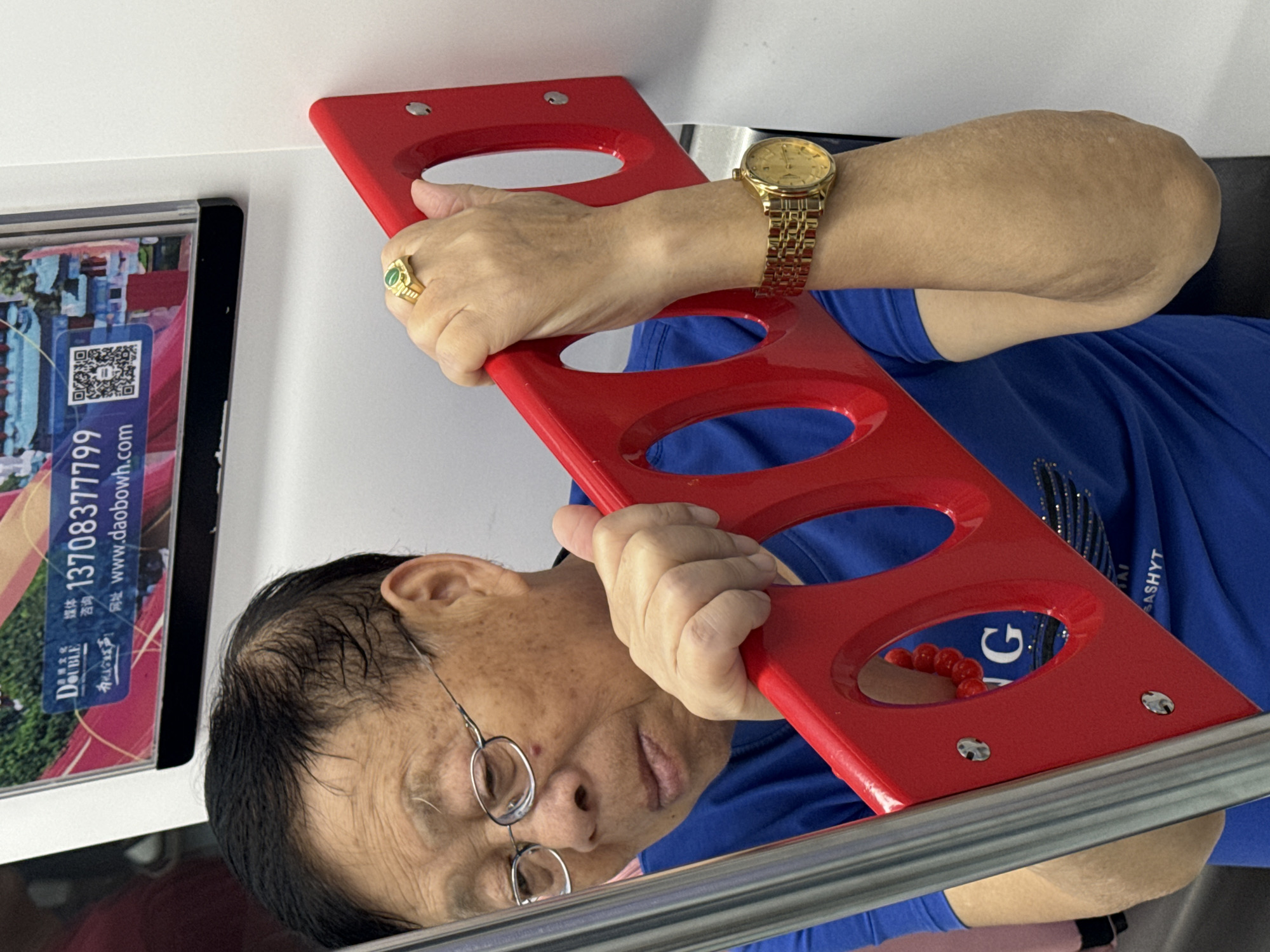 A medium shot depicts an older Asian man in a blue shirt, wearing glasses and a gold watch and ring, holding a red object with oval openings. The man's face is visible, and he appears to be looking downwards. The background includes a framed advertisement with Chinese characters and a QR code. The advertisement has the phone number 13708377799 and website www.daobowh.com. The scene appears to be indoors, likely on public transportation. The presence of Chinese characters and phone numbers suggests the location is in China, but the specific city within Yuzhong District is not identifiable from the image alone.