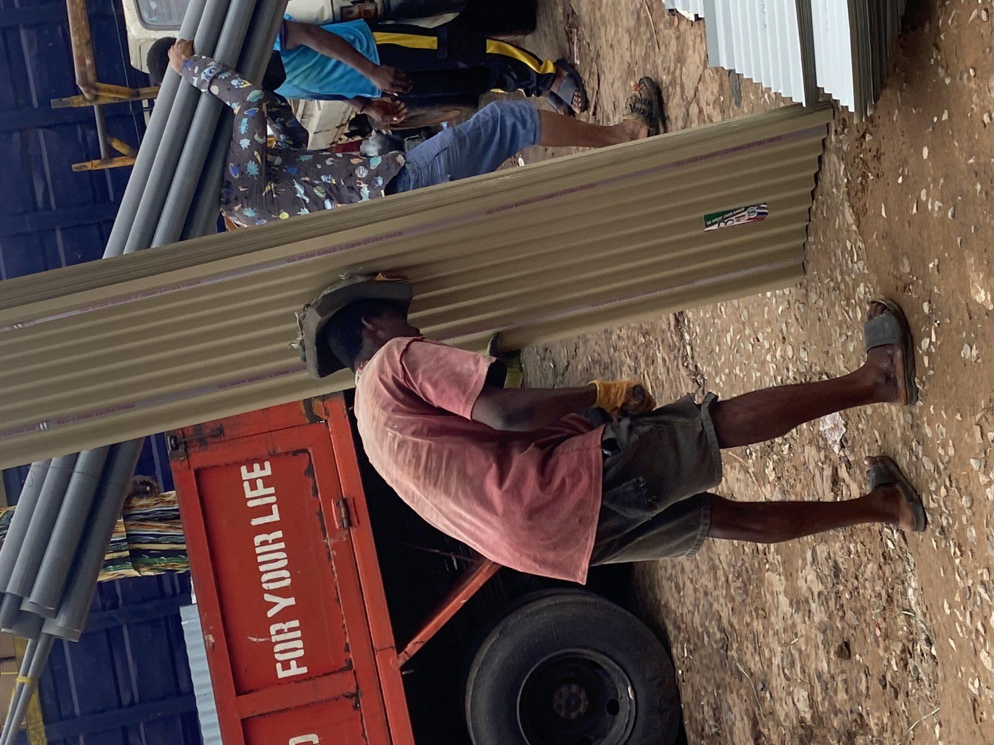 In Ogbunike, Nigeria, several individuals are engaged in the process of unloading or loading materials from a truck. A red truck with white lettering, reading "FOR YOUR LIFE," serves as the central focus of the activity. One person, wearing a red shirt and dark shorts, is bent over and appears to be supporting a large, corrugated metal sheet. Another individual, in a patterned shirt and blue shorts, is carrying a bundle of grey pipes. A third person, in a blue shirt and dark pants with yellow stripes, is also present. The environment is unpaved, with dirt and gravel on the ground. The scene suggests a construction or delivery site, with building materials like pipes and corrugated sheets being handled.