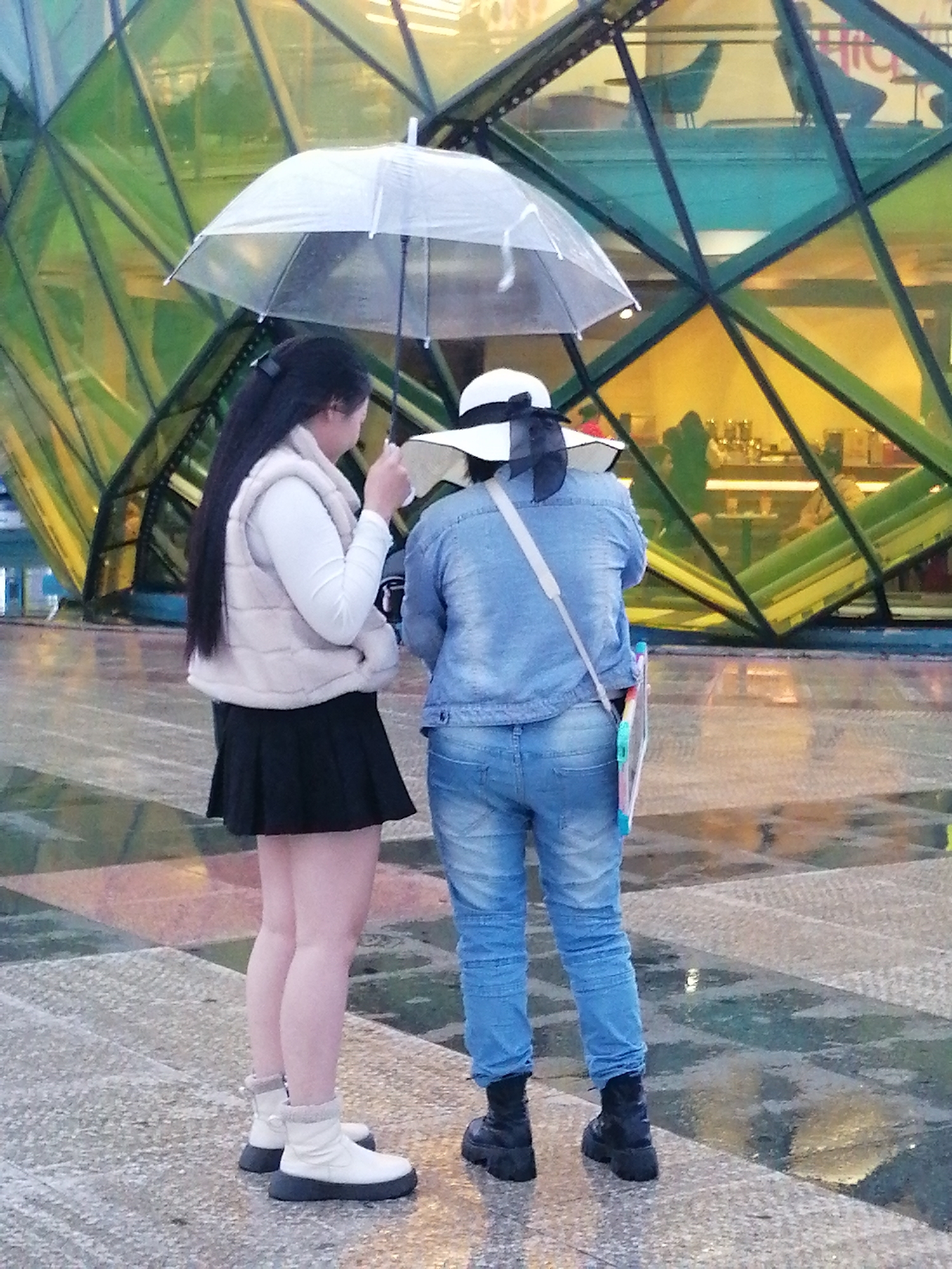 Two individuals are standing under a clear umbrella on a wet, patterned pavement in front of a building with a geometric, faceted green and yellow glass facade. The person on the left, facing away from the camera, has long black hair and is wearing a light-colored puffy vest over a white long-sleeved shirt, a black pleated skirt, and white boots with dark soles. The person on the right, also facing away, is wearing a denim jacket over a light-colored garment, blue jeans, and black platform boots. They are carrying a colorful item, possibly a drawing board or clipboard, on a strap over their shoulder. The ground is reflective due to the rain, and reflections of lights and the building are visible. The background suggests an indoor space with tables and chairs, viewed through the glass facade. This scene appears to be in Da Lat, Vietnam, possibly near a cafe or entertainment venue with a distinctive architectural design.