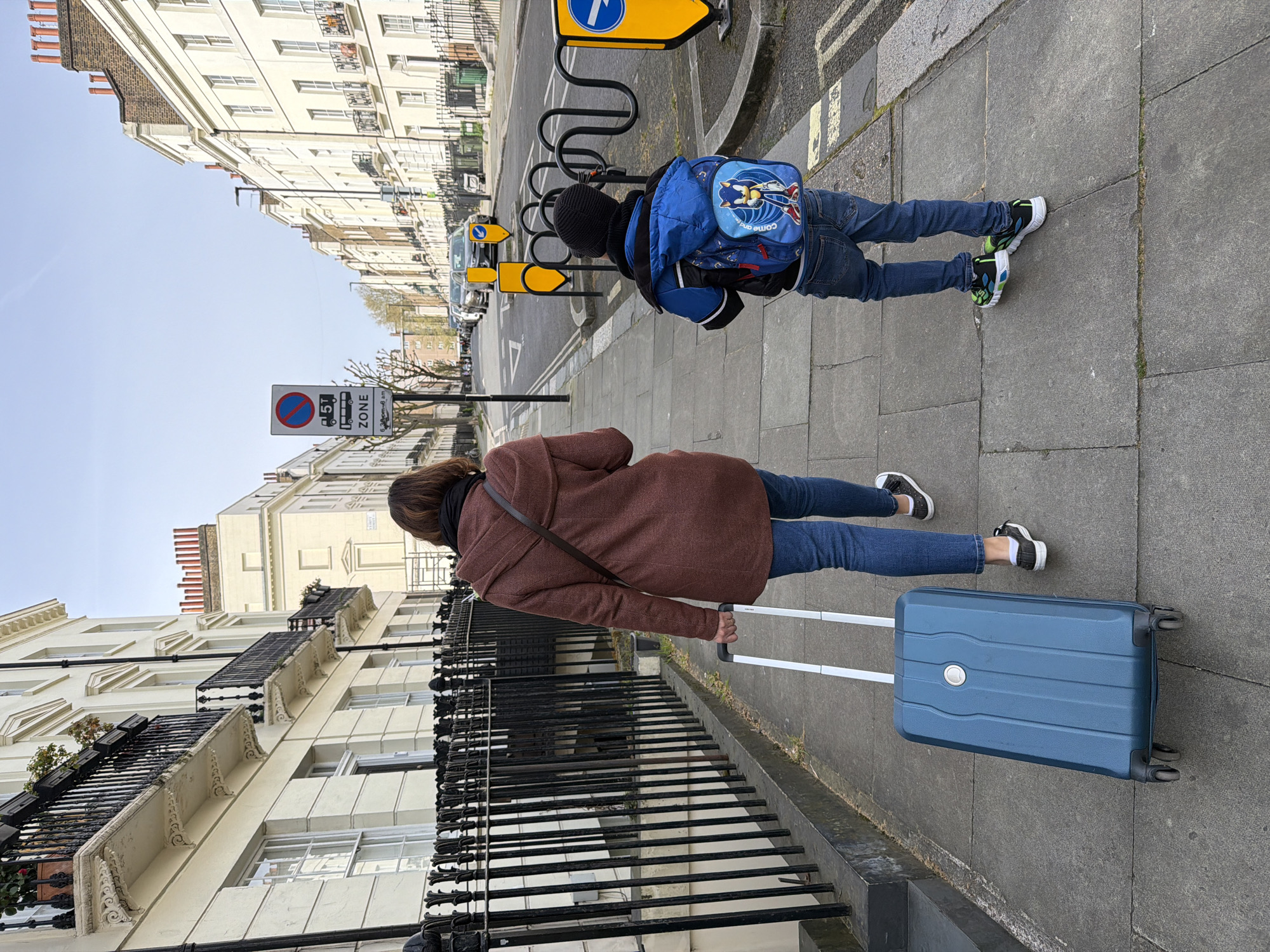 A medium-angle shot shows a woman and a child walking on a sidewalk next to a row of buildings. The woman, viewed from behind, wears a brown coat, blue jeans, and black and white shoes, and is pulling a blue suitcase. A dark brown strap crosses her shoulder. The child, also viewed from behind, wears a black beanie, a blue jacket over a blue and white backpack, jeans, and multicolored sneakers.
They are walking on a grey sidewalk next to a black metal fence. Buildings line the opposite side of the sidewalk, featuring cream-colored exteriors, windows, and decorative architectural details. A street sign is visible.
The location is likely in the city of London, United Kingdom.