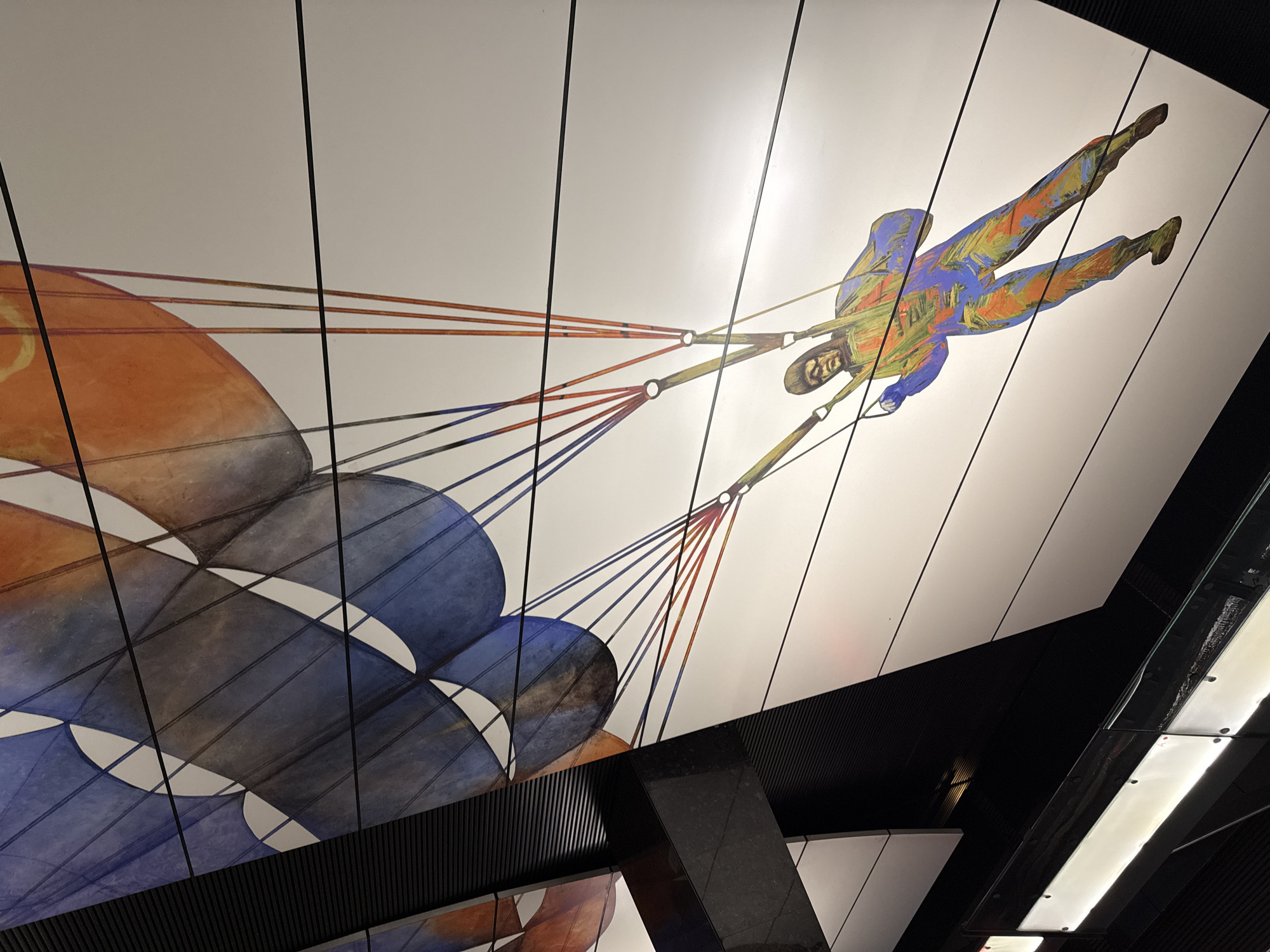 The image shows a painted ceiling mural of a parachutist in the Moscow Metro. The parachutist is depicted in mid-air, suspended beneath a partially visible parachute. The figure is painted in shades of orange, blue, and green. The parachute is depicted in shades of orange, blue, grey, and white. The ceiling is segmented, with black architectural features and lighting fixtures visible in the corners and edges of the frame. The mural's subject matter and style suggest it is part of the Mayakovskaya Station's art deco design.