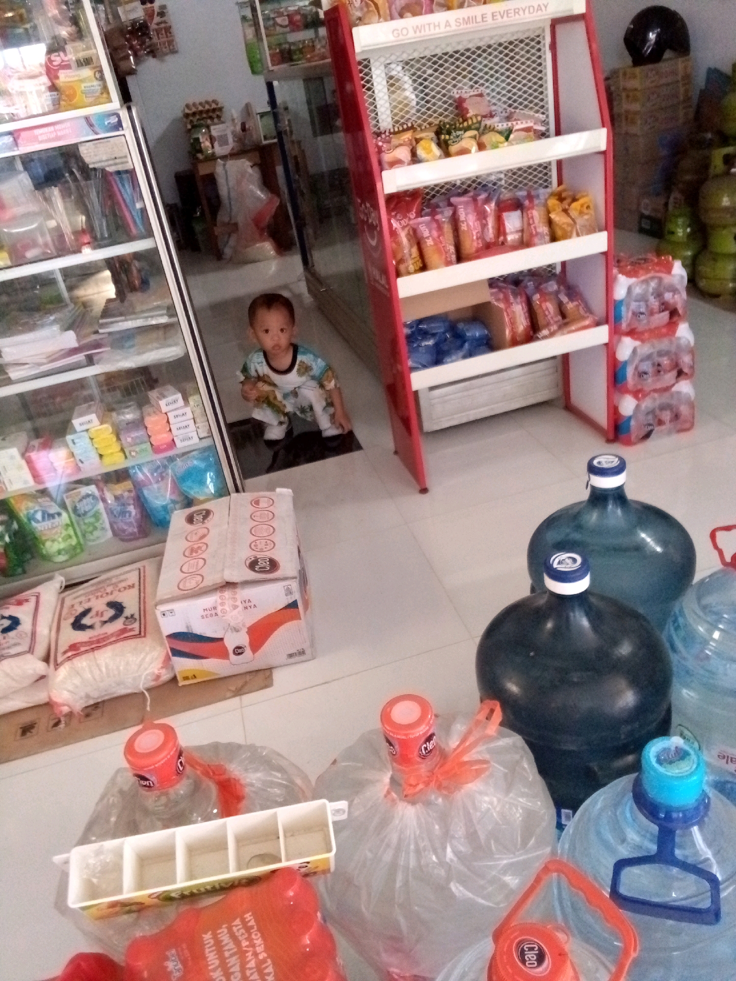 The image depicts the interior of a small convenience store, likely in Simo, Indonesia, as indicated by the store's inventory which includes many common Indonesian household goods. A young child, appearing to be under two years old, is crouched on the floor near a display shelf filled with packaged snacks. The child is wearing a light-colored outfit with a colorful pattern.
In the foreground and to the right, several large, blue-tinted water jugs are visible. Some are covered with plastic bags. A white plastic dispenser with multiple compartments is positioned on top of one of these covered jugs. Various other packaged goods, including rice bags and boxed items, are scattered on the floor. The shelves behind the child hold a variety of items, including bottles, boxes, and what appear to be cleaning supplies.
The store appears to be well-stocked with a mix of food items, beverages, and household necessities. The lighting suggests it is daytime, and the overall environment is that of a typical local shop.