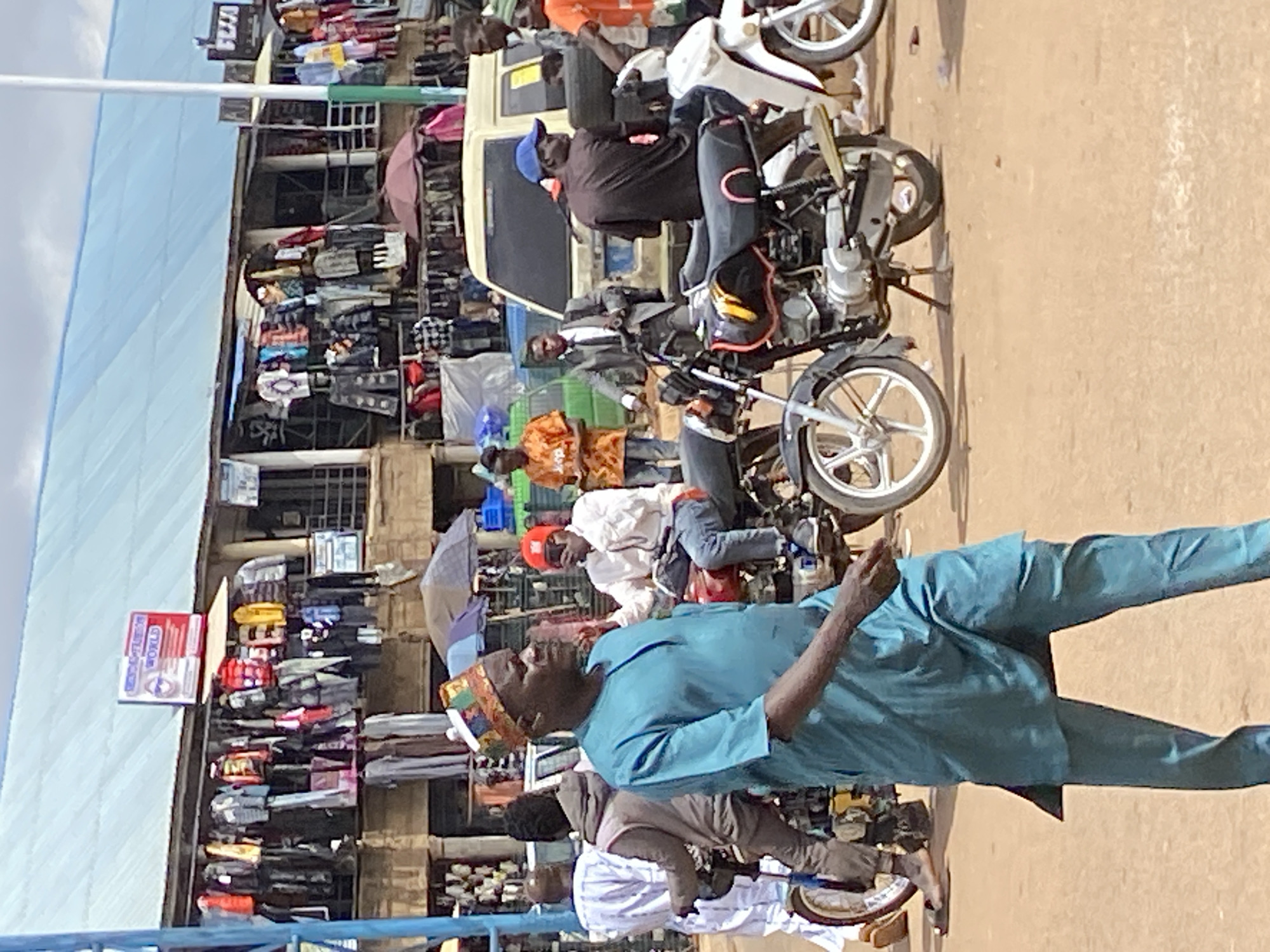 In Bukuru, Nigeria, a bustling marketplace is depicted on a bright, overcast day. A man in a turquoise traditional outfit and a colorful kufi hat walks towards the viewer in the foreground, his body angled to the right. Behind him, a crowd of people are engaged in various activities. Several motorcycles are parked, with some individuals seated on them. A light-colored van is visible behind the motorcycles. The background features market stalls overflowing with clothing, ranging from jackets to shirts and dresses, all hanging on display. A blue awning stretches across the top of the stalls. The ground is a dusty, sandy surface typical of an outdoor market.