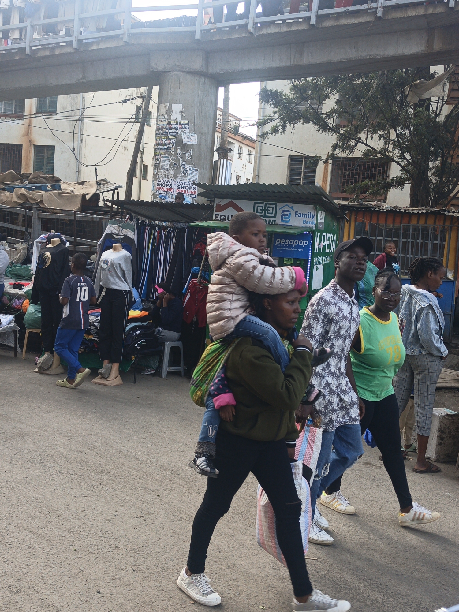 The image captures a vibrant street scene in Nairobi, Kenya. A woman carries a child on her shoulders as she walks along a busy street. The child wears a pink hat, a puffy jacket, and jeans, while the woman is dressed in a green jacket, black pants, and white sneakers and carries a striped bag. Next to them, a man with a black cap is wearing a patterned shirt and torn jeans. To his right, a woman with glasses is dressed in a green top and black pants.
Several other pedestrians walk on the street, passing by open-air market stalls lined with clothing and various goods.
A building stands in the background, a concrete structure supported by pillars with posters affixed to them. The area is crowded with people, merchandise, and the infrastructure of daily life in an urban setting.
There are signs of local businesses and advertising, including signs for "Family Bank" and "PESA."