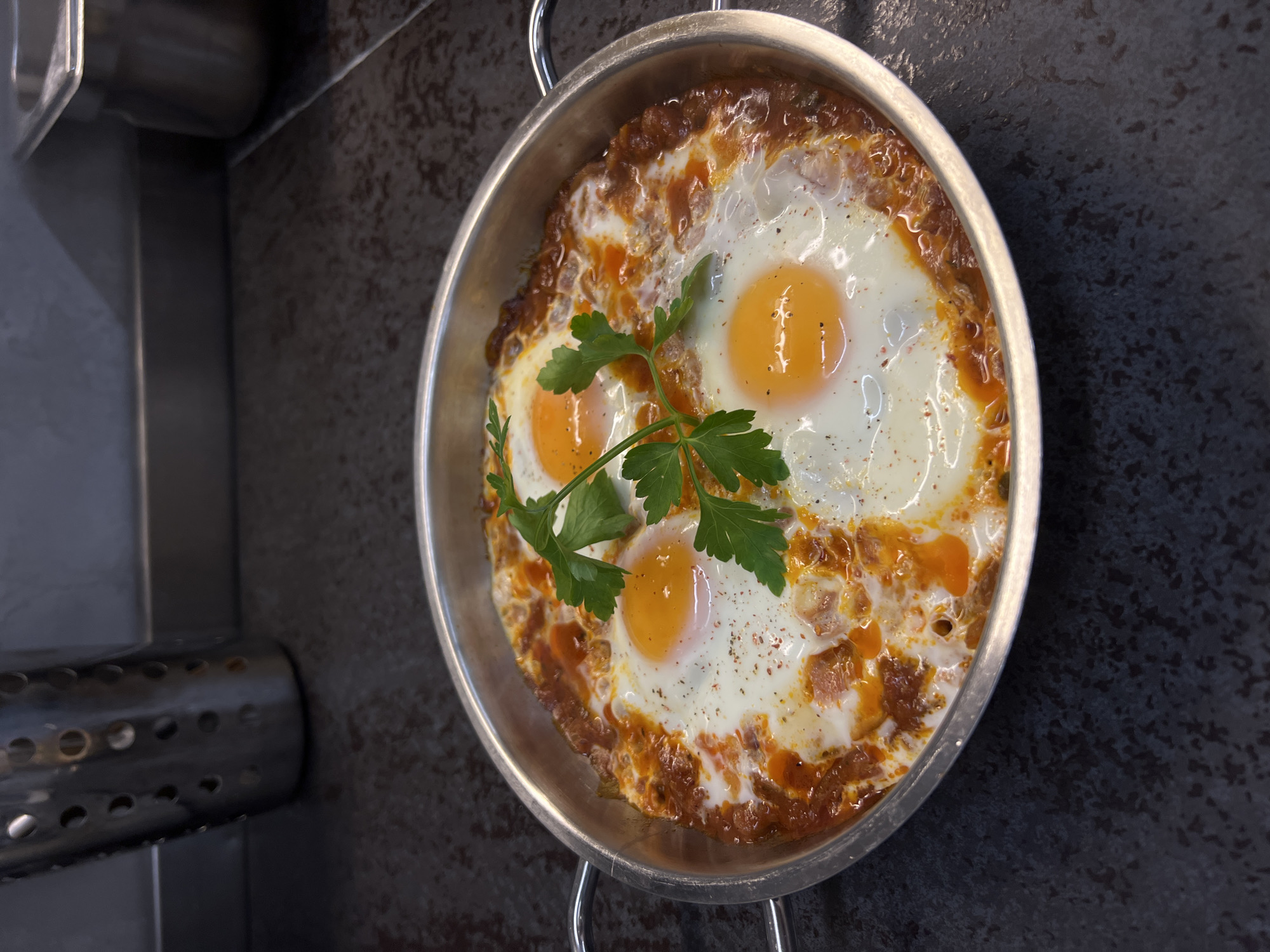 The image shows a silver-colored cooking pan with two loop handles, containing a dish of three sunny-side-up eggs nestled in a tomato-based sauce, garnished with a sprig of parsley. The pan sits on a dark, textured surface, and a stainless steel apparatus with circular perforations is visible to the left. The location of the image is not discernible, and information about people, actions, or interactions is not present. The location is not identifiable as Budva, Montenegro.