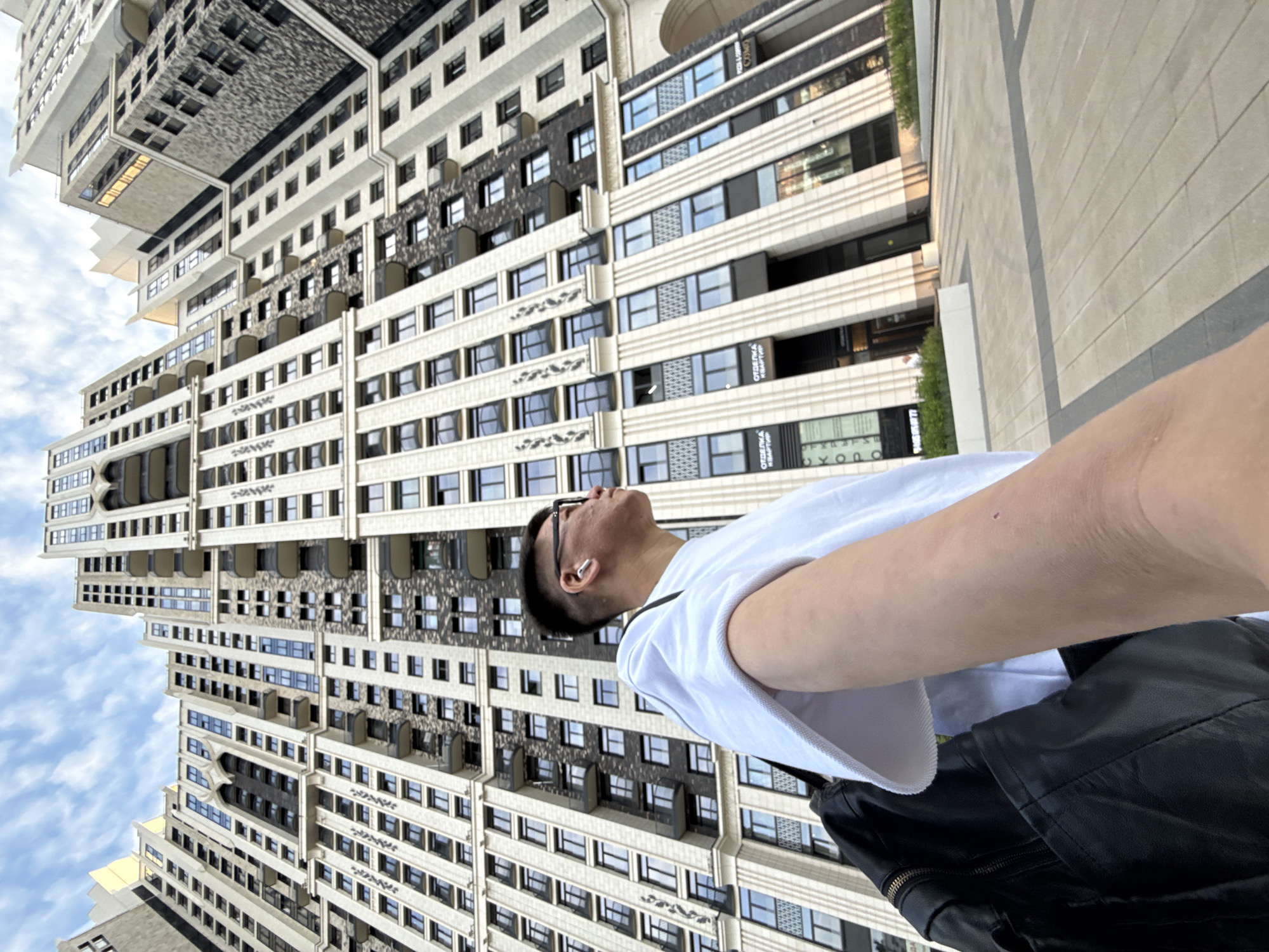 An individual, with short dark hair and glasses, is seen from a low angle, looking upwards and to the right. They are wearing a white t-shirt and a black backpack. In the background, a tall, modern, multi-story building with a light-colored facade and dark windows dominates the scene. The building has balconies and intricate architectural details. The sky above is partly cloudy. The ground in front of the building is paved with large light-colored tiles. Signs with Cyrillic text are visible on the building's lower levels. The overall impression is an urban setting with a focus on contemporary architecture.