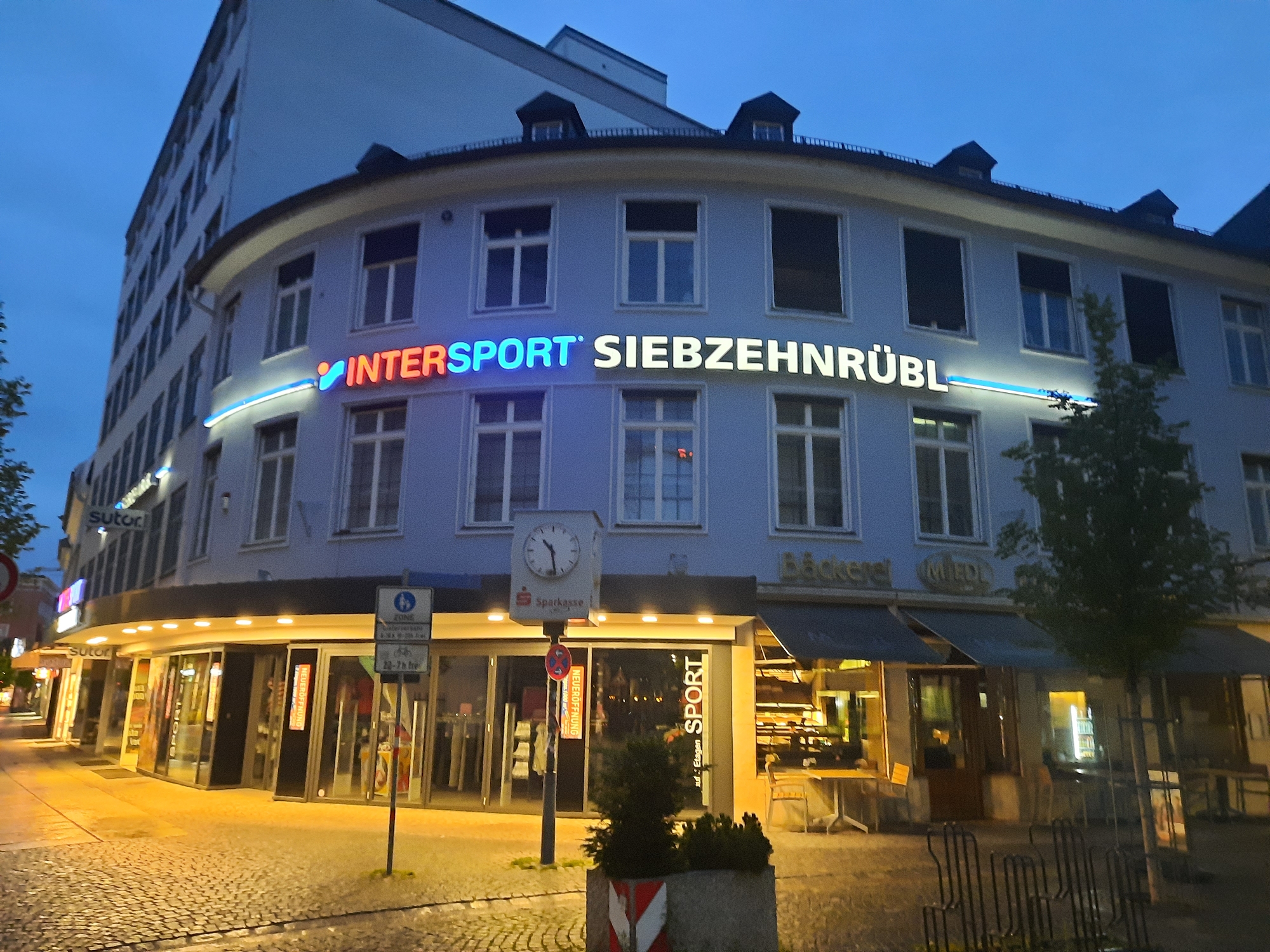 The image depicts the exterior of a curved building at dusk on a street in Rosenheim, Germany. The building houses several businesses, most notably an "Intersport" store with illuminated signage. To the left of Intersport, the sign "sutor" is visible. To the right of Intersport, there is a bakery identified by the signage "Bäckerei" and "MEI" which is likely part of a larger name. A clock tower with the "Sparkasse" logo is mounted on the building above street level. The street in front of the businesses is paved with cobblestones and is lit by streetlights. There are no people clearly visible in the image, though there may be individuals inside the stores. The overall scene suggests a commercial area in a German city at the end of the day.