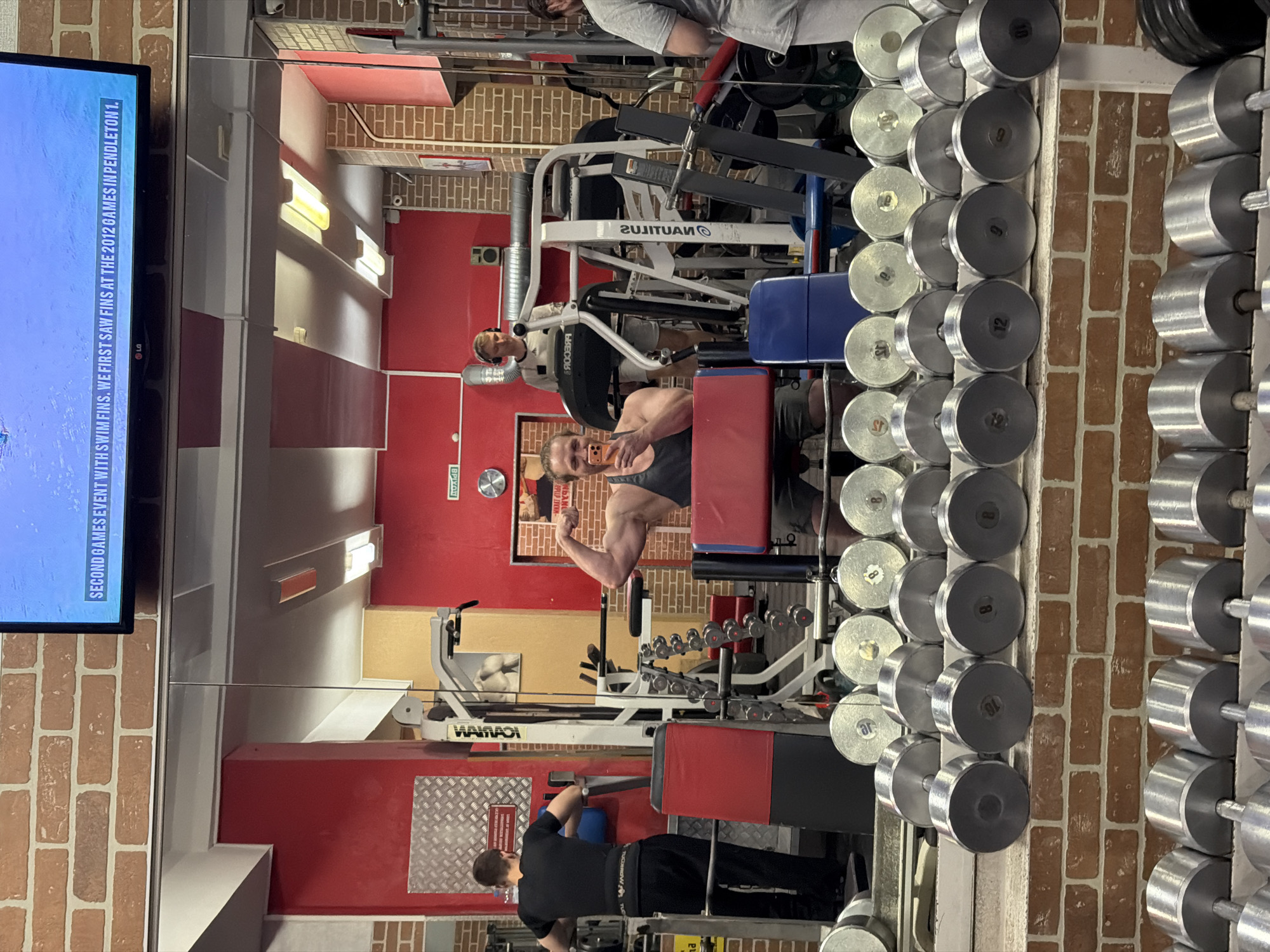 Here's a description of the image:
**Location:** The image appears to be taken inside a gym with red and brick-textured walls. The specific location cannot be determined from the visual data provided.
**People:** There are at least three people visible in the image.
* One man is flexing his bicep while taking a selfie in the mirror.
* Another man is visible behind the selfie taker and appears to be using a machine.
* A third man is seen in the lower part of the image, working out on a machine.
**Subjects:** Gym equipment, including various machines, a dumbbell rack, and dumbbells, are present. There is also a television screen showing sports programming.
**Objects:** Dumbbells of various weights, Nautilus workout equipment, a television.
**Actions:**
* One man is taking a selfie while flexing.
* Other people in the gym are engaged in various workout activities.
**Interactions:** The man taking the selfie is the primary subject of the image and appears to be the person taking the photo. The other individuals are present and working out in the background.
**Environment:** The gym has a red and brick interior design with standard gym equipment. The dumbbells are racked, and the machines are arranged in a functional layout. There is also a television screen playing sports programming.
