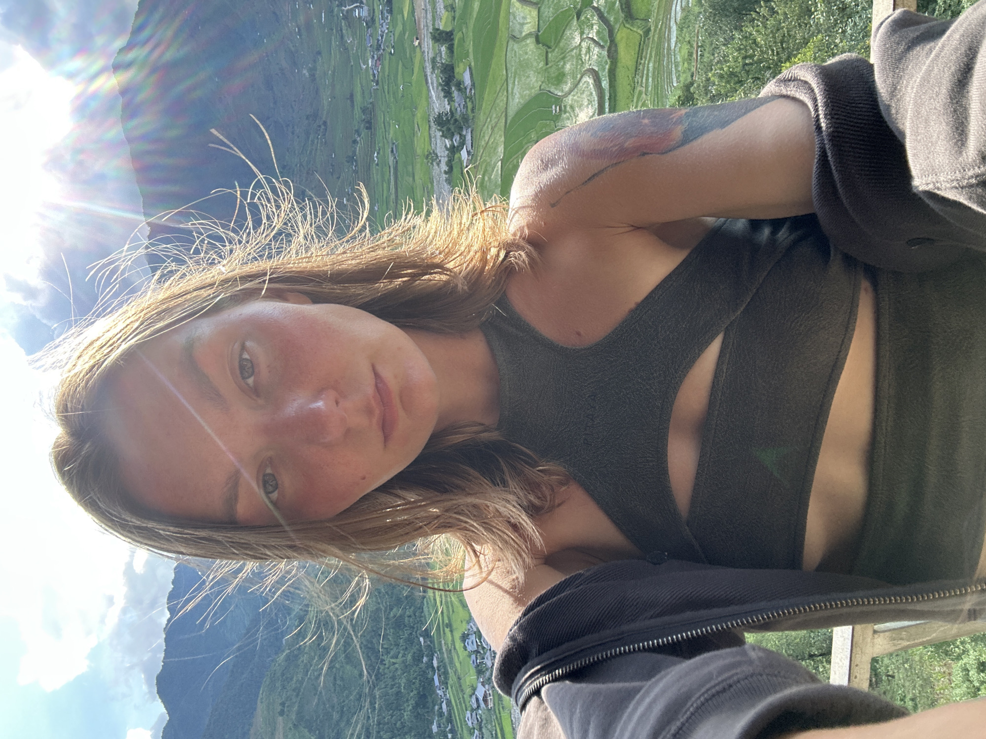 A female subject, positioned centrally, takes a selfie outdoors against a panoramic mountainous backdrop, identifiable as Tu Le, Vietnam. Her light brown, shoulder-length hair is illuminated by strong backlighting and appears wind-swept, with numerous individual strands visible. Her face is directed towards the camera with a neutral expression, exhibiting visible freckles across her nose and cheeks, which are flushed with a reddish tone. Her eyes are light in color. She wears a dark gray, halter-style cut-out top with an exposed midriff section below the bust. A dark gray zip-up hoodie or jacket is draped over her shoulders and arms. A multi-colored, abstract tattoo is visible on her upper left arm. The background features extensive, verdant terraced rice fields covering the slopes of multiple mountains, interspersed with a village containing numerous small houses. The sky is partly cloudy, and intense sunlight emanates from the upper right quadrant, producing significant lens flare and distinct rainbow-like chromatic aberration. A light-colored railing is partially visible in the bottom left corner of the frame.