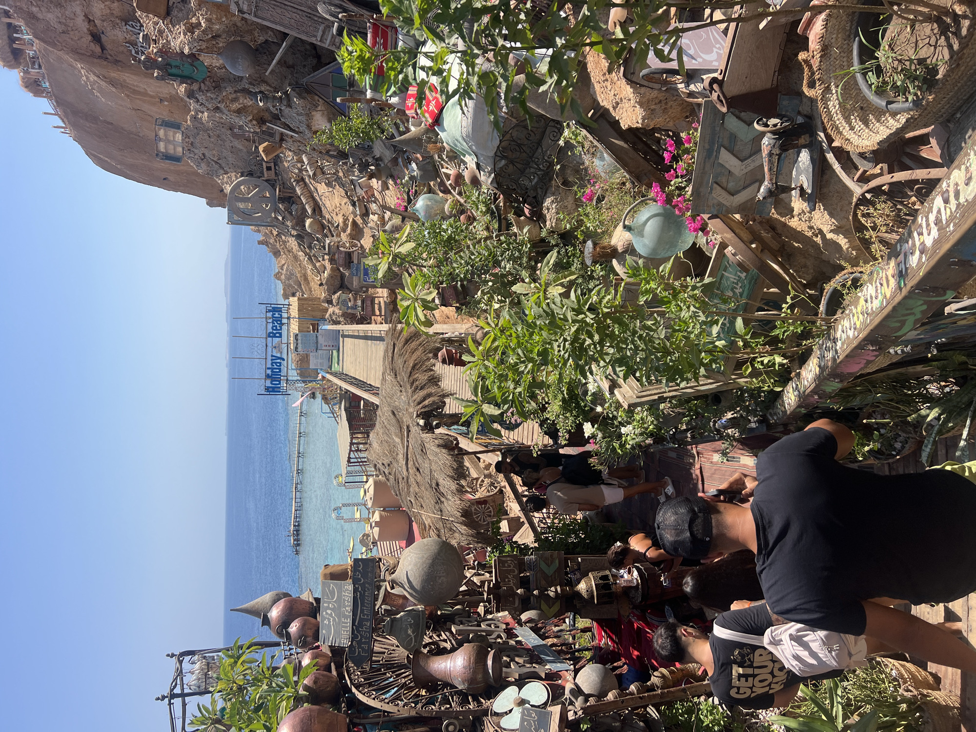 The image depicts a vibrant outdoor market or cafe area built into a rocky hillside overlooking the Red Sea in Sharm El Sheikh, Egypt. A wooden walkway guides visitors down a steep path lined with an eclectic assortment of decorative items, including large clay pots, metal sculptures, antique machinery, baskets, and plants. In the background, the turquoise waters of the sea are visible, with a pier extending out and a "Holiday Beach" sign overhead. Several people are walking down the path, some appearing to be tourists. The overall atmosphere is bohemian and rustic, with a strong sense of place and cultural character.