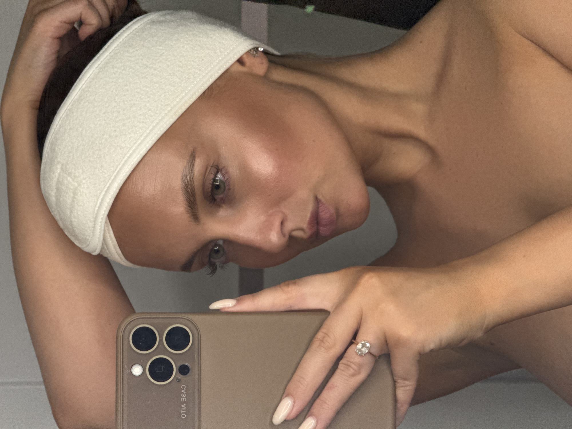 The image shows a woman taking a selfie in what appears to be a bathroom setting. She is wearing a white terry cloth headband and is holding a brown smartphone with a multiple lens camera array. Her right hand is visible, and she wears a ring with a large clear stone on her ring finger. Her skin has a dewy appearance, possibly indicating the presence of skincare products. The background is indistinct, with a light-colored wall visible. There is no information in the image that places the location in the city of Chaikovsky, Russia, or that mentions any subjects or actions in that location.