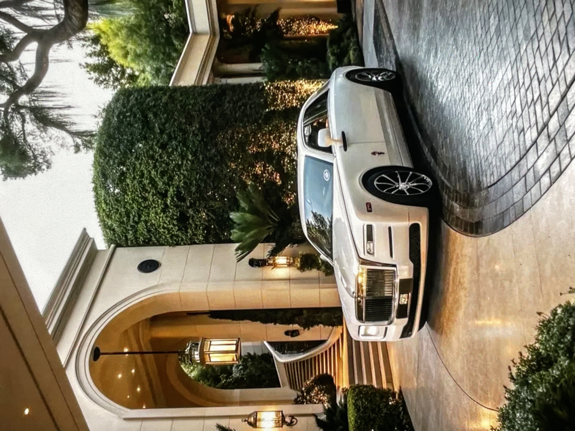 The image displays a white Rolls-Royce sedan parked on a paved driveway in front of a grand entrance of a mansion. The architecture of the mansion features a large arched entryway with ornate lighting fixtures and a sweeping staircase leading upwards. Lush green landscaping, including manicured hedges and trees, surrounds the property. The driveway itself is a combination of smooth concrete and patterned pavers, reflecting the ambient light. The overall scene suggests a luxurious and affluent setting. The provided context of the image being from Yalga, Russia, is not visually verifiable from the image itself.