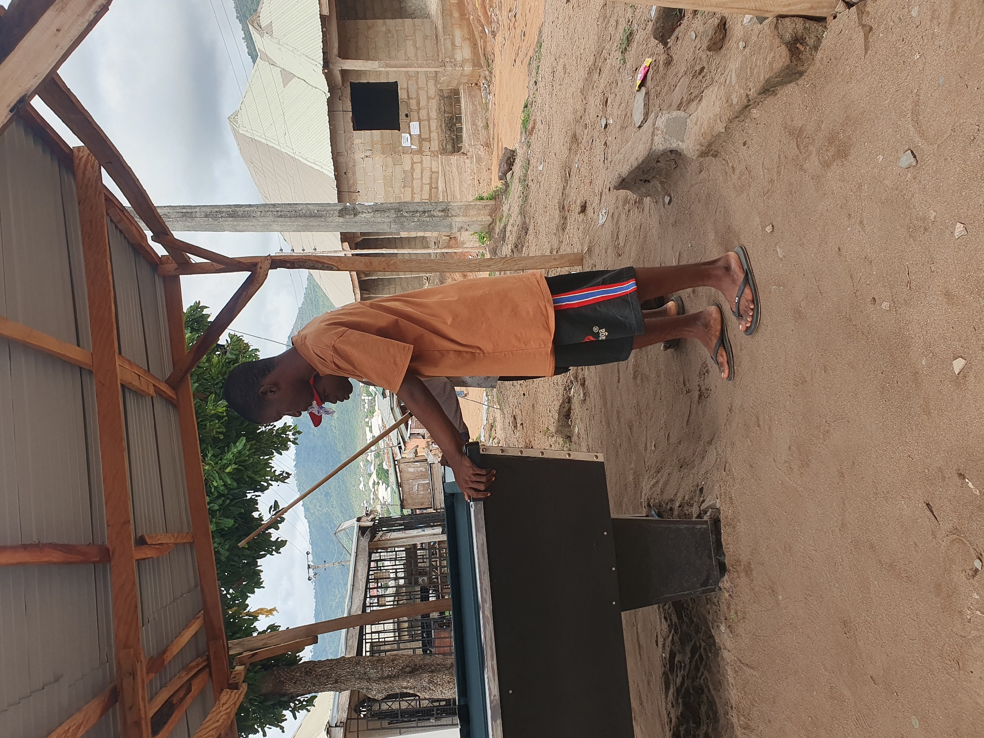 A young man in an orange shirt, black shorts with red and blue stripes, and flip-flops stands beside a pool table under a wooden structure with a corrugated metal roof in Lokoja, Nigeria. He is leaning over the pool table, seemingly inspecting it. The ground is sandy. In the background, there are buildings, including an unfinished concrete structure with an open window. Trees and distant hills are also visible.