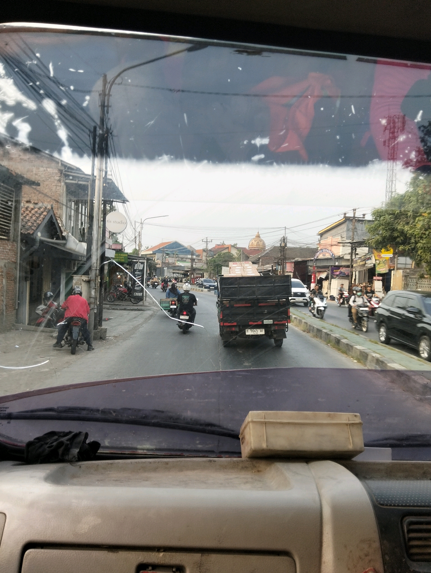 The image depicts a street scene in Semarang, Indonesia, as viewed from the interior of a vehicle, likely looking through the windshield. The street is paved and shows moderate traffic, predominantly motorcycles. In the foreground, a light-colored plastic box is placed on the dashboard.
On the left side of the street, there are buildings with traditional Indonesian architecture, including tiled roofs and brick facades. A person wearing a red jacket is seated on a motorcycle, facing away from the camera. Further down the street, there is a black truck carrying boxes, also moving away from the viewer. A white car is seen behind the truck.
The right side of the street features more buildings, some with signs and awnings. Several motorcycles with riders are in motion. One rider is wearing a white helmet, another a dark helmet, and a third appears to be wearing a face mask. In the distance, more buildings and trees are visible, suggesting a developed urban area. The sky is overcast. Reflections on the windshield obscure some details of the scene.