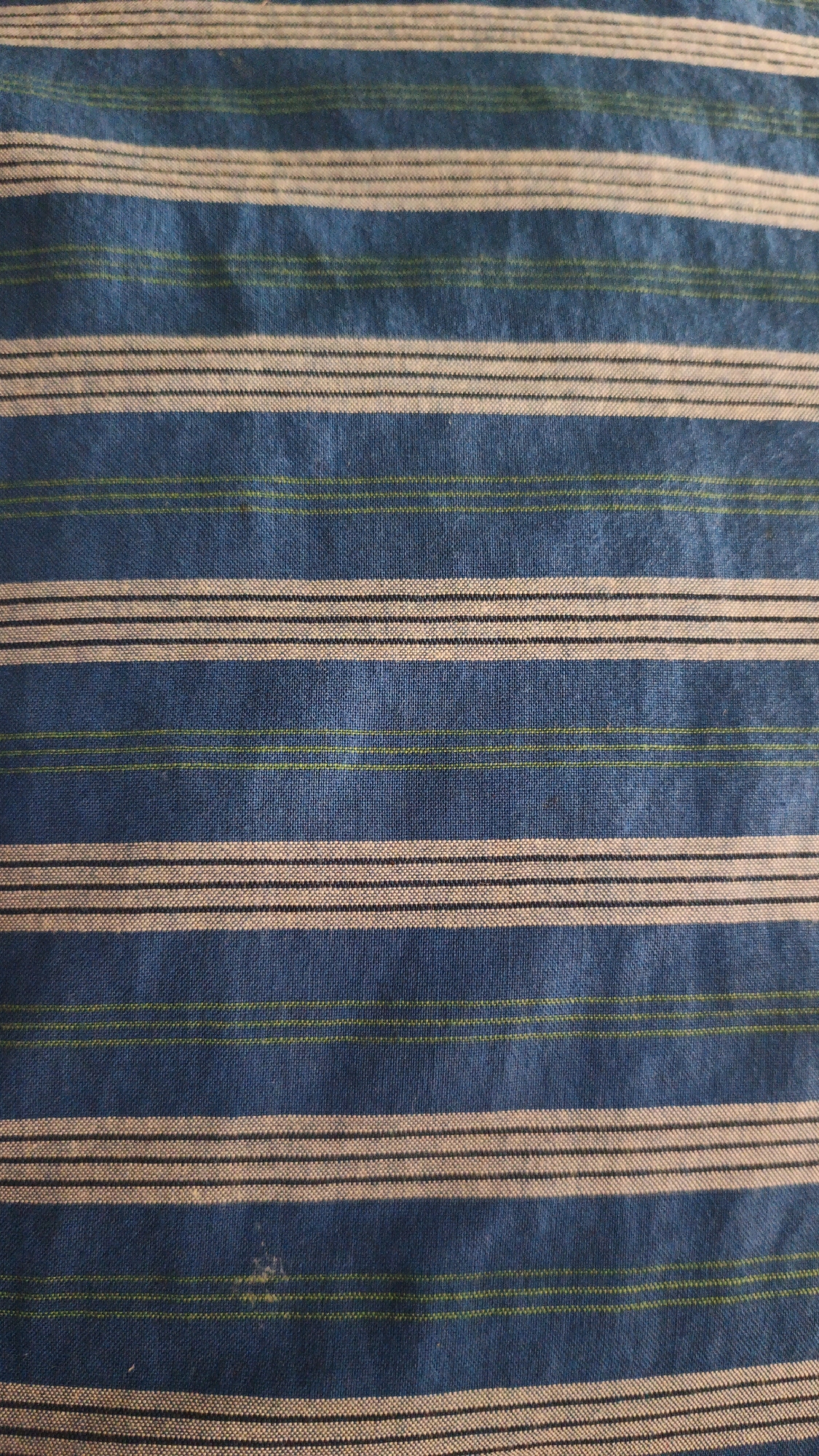 The image shows a close-up view of a striped fabric. The stripes are horizontal and vary in thickness and color. The dominant color of the fabric is a medium blue. There are wider stripes of a lighter color, possibly off-white or beige, and thinner stripes of black and a light green. The texture of the fabric appears to be woven. There is a small stain of an indeterminate color on the fabric in the lower central area. The image is a flat lay of the fabric and there is no context to determine its location in Santing, Indonesia.