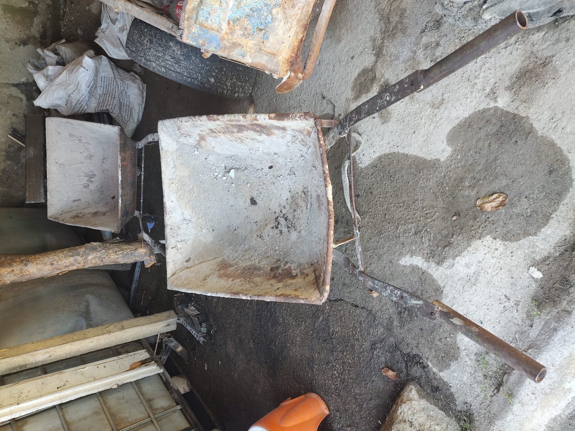 The image depicts a cluttered outdoor storage area, likely a yard or shed, in Kochmar, Bulgaria. The foreground is dominated by a rusty, used wheelbarrow with a large, worn tray filled with dust and debris. Its metal handles are rusted and extend towards the viewer. Behind this wheelbarrow, another, similar wheelbarrow is partially visible. To the right, a section of a rusty, blue-painted metal object, possibly another piece of equipment or a damaged container, is present. In the background, several large, white woven bags, likely containing building materials or soil, are stacked. A worn-out tire is also visible. The ground appears to be a mixture of concrete and dirt, with some puddles of water and scattered leaves. Various construction-related items, including wooden planks and what looks like a broken jack stand, are strewn about, contributing to the overall impression of disarray. The scene is lit by natural light, casting shadows on the ground. There are no people present in the image.