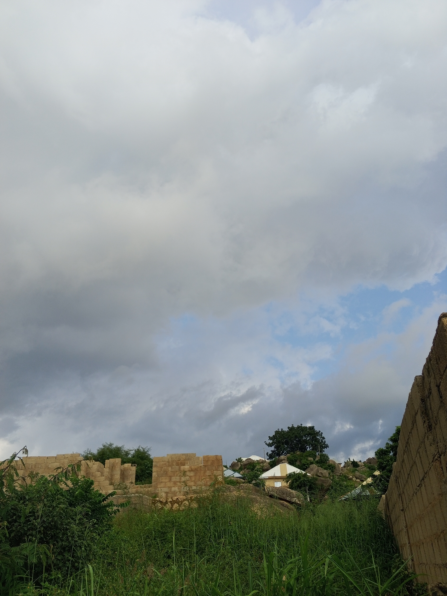 The image depicts a cloudy sky over a rocky, overgrown landscape in Kabong, Nigeria. In the foreground, tall green grass and foliage dominate, with some of the blades reaching into the lower frame. To the left, a partially constructed building made of light brown bricks is visible, with several openings suggesting it is either unfinished or in ruins. Lush green bushes surround the brick structure, partially obscuring it.
Further into the scene, the terrain becomes more rugged with large rocks and boulders interspersed with more vegetation. Several houses with light-colored roofs are nestled among the rocks on a hillside, some appearing to be in better condition than the brick structure in the foreground. A mature, dark green tree stands prominently on the hill.
On the right side of the image, a rough, unfinished concrete wall extends upwards, creating a stark contrast with the natural elements and the brick building. The sky is filled with a mix of light and dark gray clouds, with patches of blue sky peeking through. The overall impression is one of a somewhat undeveloped or rural area, possibly during an overcast or pre-stormy day. No people or specific activities are visible. The time of day is indeterminate due to the cloud cover, but the lighting suggests it is daytime.