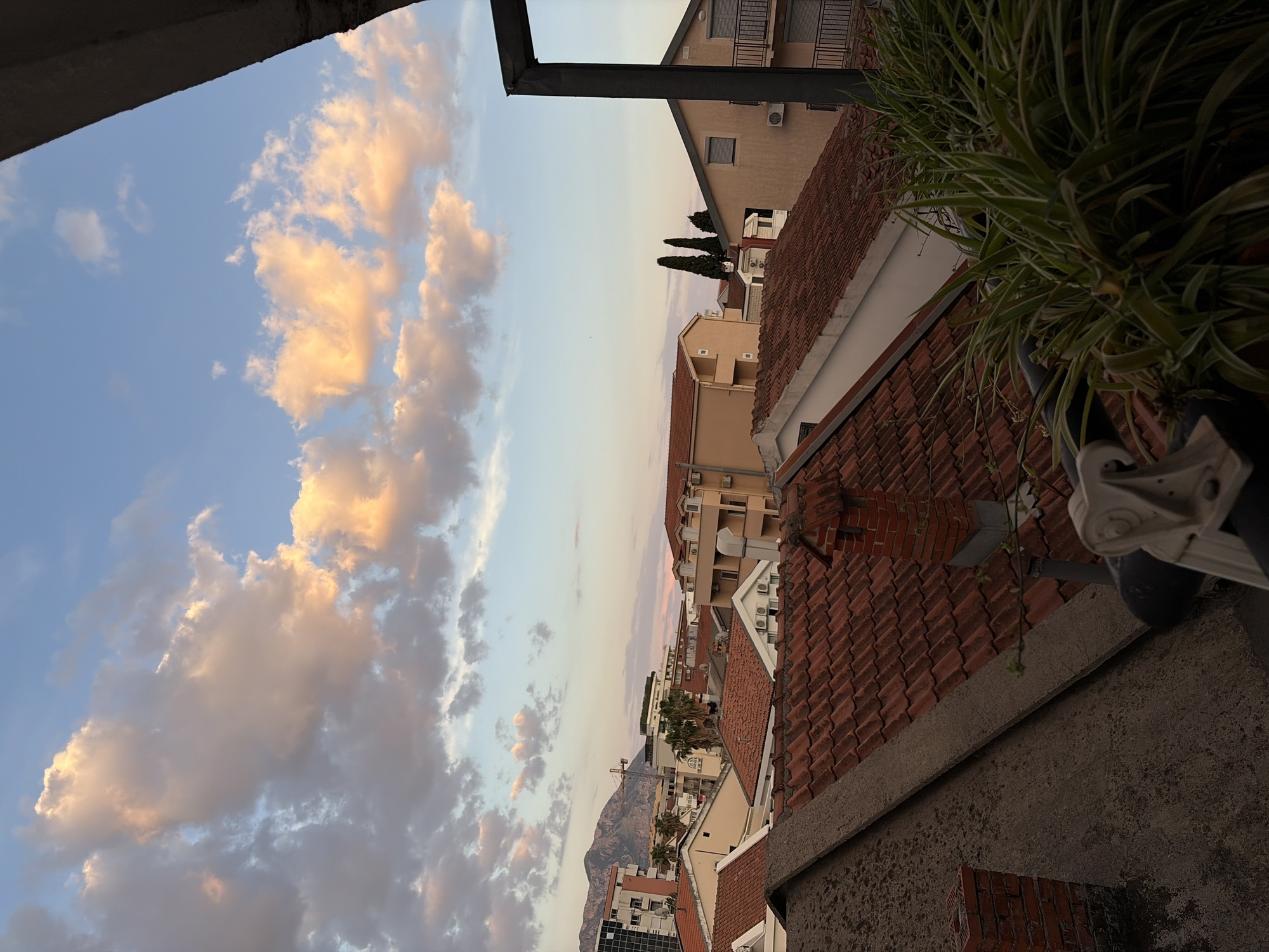 A high-angle, landscape-oriented photograph captures a scene in Budva, Montenegro, at what appears to be either sunrise or sunset given the coloration of the sky.
The photograph is framed by architectural elements at the top, suggesting it was taken from a window or balcony. To the left, the sky is filled with clouds that are tinged with golden light, indicating the presence of the sun below the horizon. The sky transitions to a softer, lighter tone towards the center of the frame.
The middle ground is occupied by buildings with red-tiled roofs, characteristic of Mediterranean architecture. Some buildings are taller and painted in light colors. A mountain is visible on the horizon.
In the right foreground, part of a building’s structure is visible. A potted plant with long, thin leaves is present.