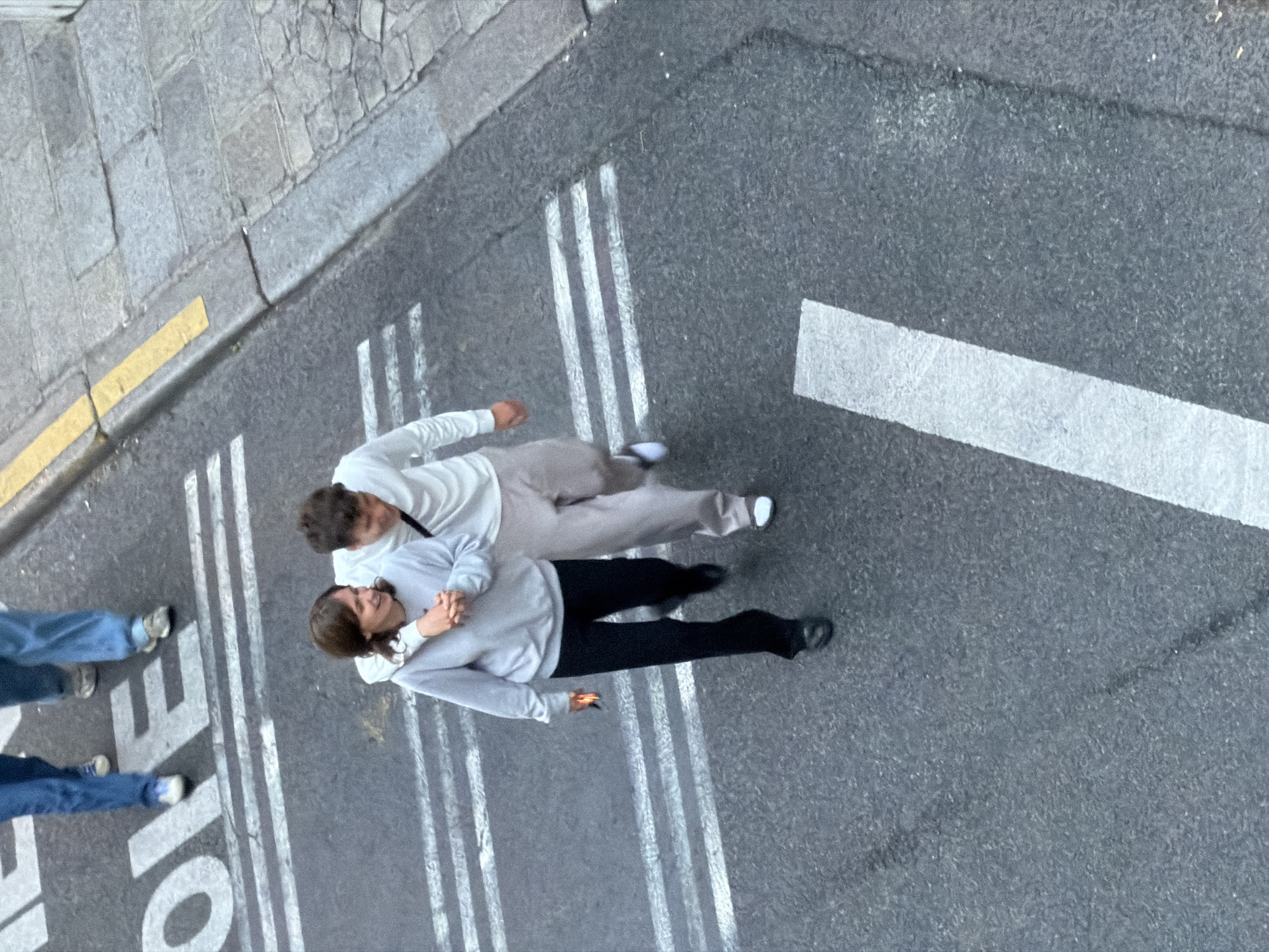 The image shows two people embracing while standing in a pedestrian crosswalk on a street in Paris, France. One person wears a white sweatshirt and beige pants, while the other wears a light gray sweatshirt and black pants. The crosswalk is marked with white lines on an asphalt road. The adjacent sidewalk is paved with rectangular stones. A yellow line marks the edge of the sidewalk. Two additional people are walking on the crosswalk. A traffic sign that reads "Ecole" is marked on the crosswalk.