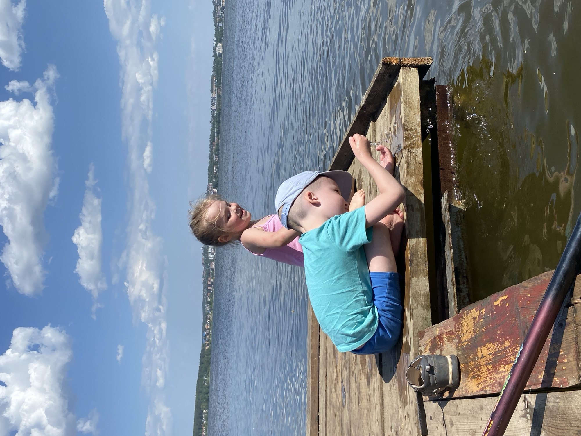Two young children, a boy and a girl, are sitting on a wooden dock or pier. The boy, wearing a blue shirt, shorts, and a cap, is dangling something into the water. The girl, wearing a pink top, is looking at the water and the boy. The dock extends into the body of water, which appears to be a lake or river. The background shows a cityscape with buildings, presumably the city of Votkinsk, Russia. The sky is blue with some white clouds. A child's shoe sits on the dock near the boy.