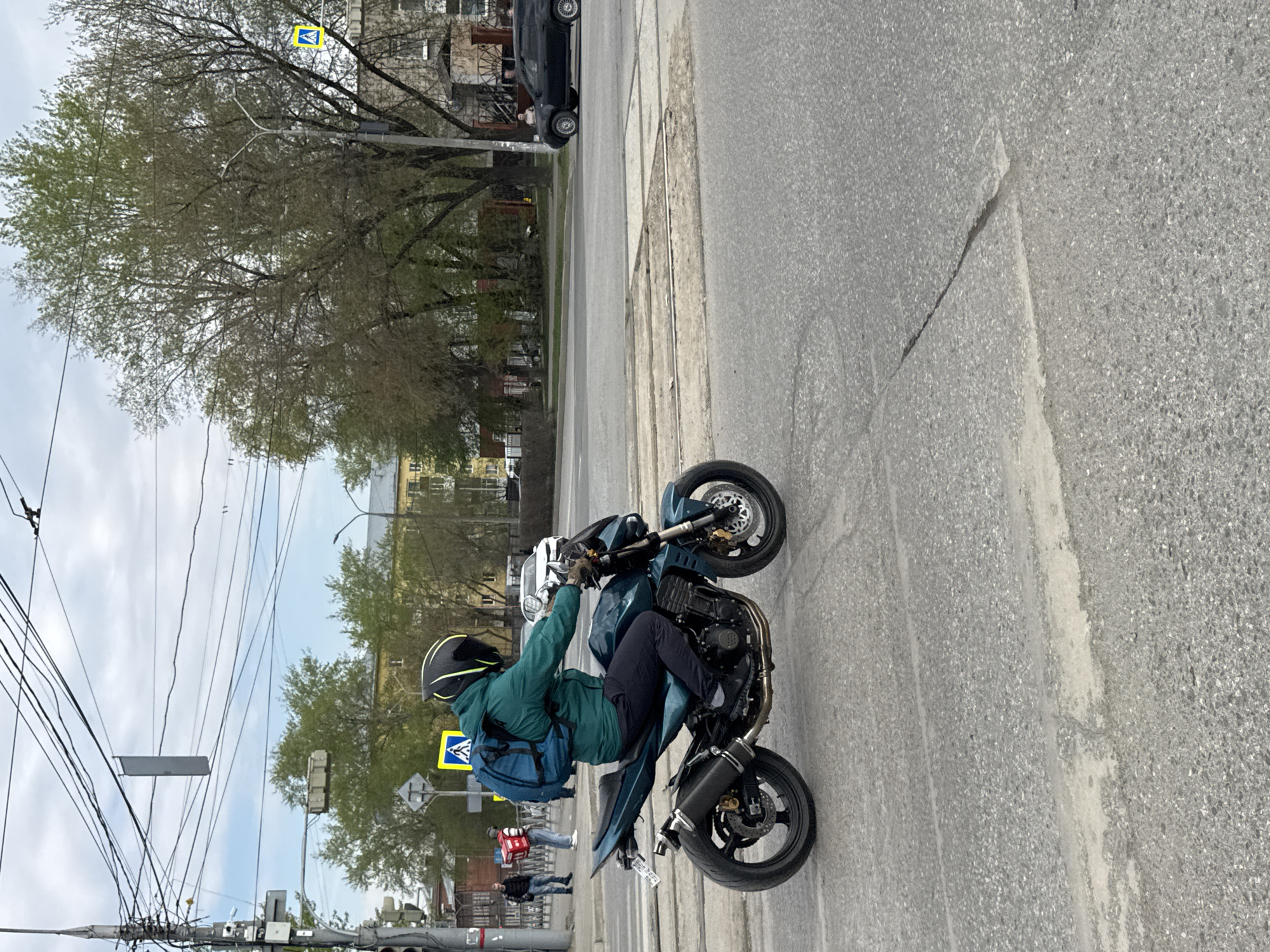 A man performs a motorcycle wheelie on a city street in Perm, Russia. The motorcyclist wears a black helmet with yellow accents, a green jacket, and a blue backpack. The motorcycle is teal and black. To the left, a pedestrian crossing sign is visible, with people standing on the sidewalk. Power lines are overhead. A building is visible in the background, as well as a parked black car. The street has visible wear and tear.