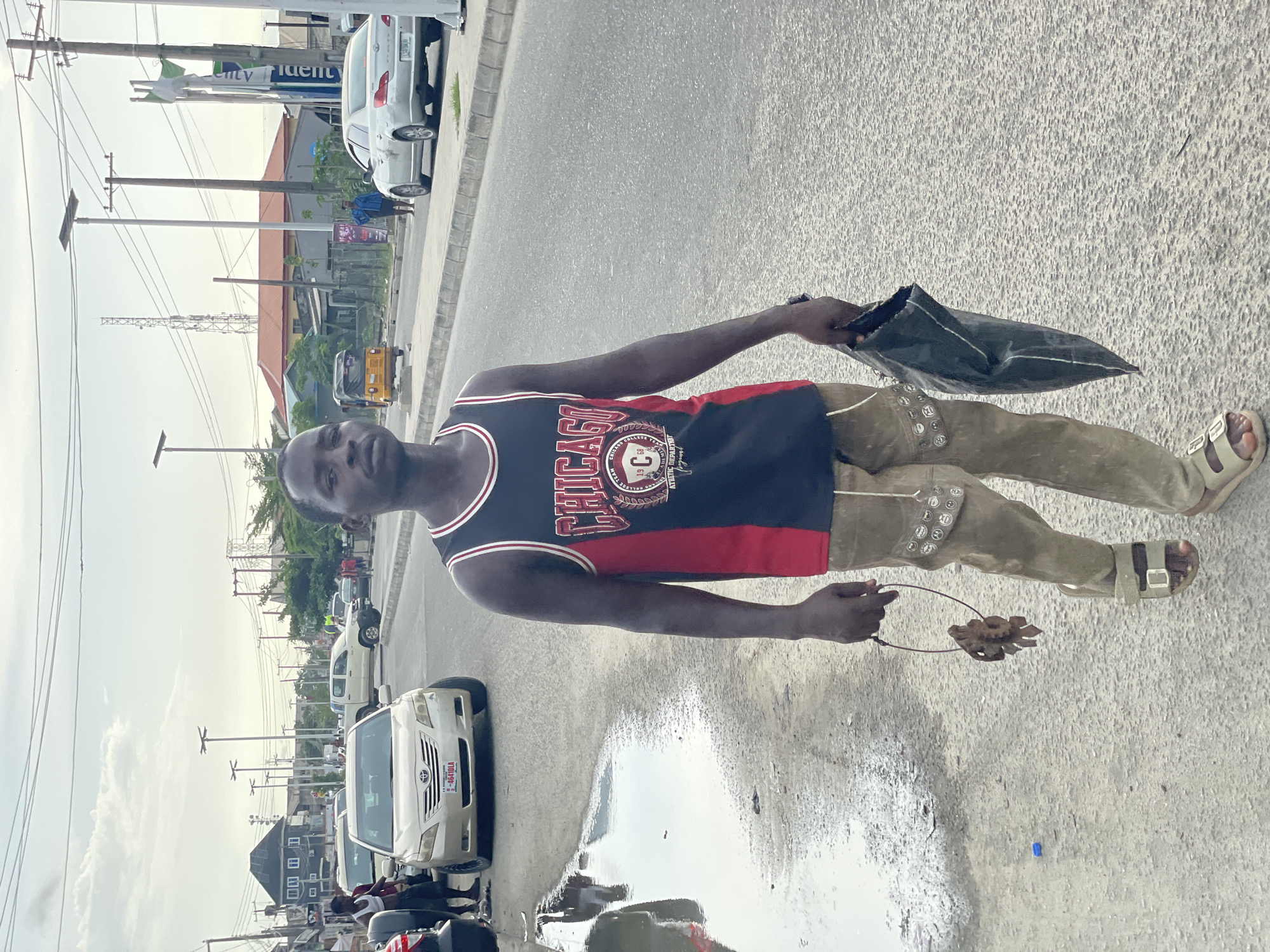A young Black male is centrally positioned on an asphalt road in an urban or suburban setting in Ado, Nigeria. He stands looking directly at the viewer. He is wearing a black and red Chicago Bulls basketball jersey, tan-colored pants embellished with metallic circular studs on the left thigh, and light-colored, open-toed sandals. In his right hand, he holds a crinkled black bag, and in his left hand, he holds a small, decorative object, possibly a keyring or a small craft, shaped like a flower or gear, attached to a thin wire loop.
The background reveals a bustling street scene with numerous utility poles and an intricate network of overhead power lines. Vehicles are visible, including a white Toyota sedan with the license plate "ABJ 441ULU" parked behind the subject, and other cars further down the road. A yellow tricycle, commonly known as a Keke Napep, is also partially visible. Along the street, there are various buildings, including residential homes and commercial structures, one of which has a partially visible sign displaying "city" and "udent." In the distant background, a tall antenna or telecommunication tower can be seen against the sky.
The road surface in the foreground and around the subject is mostly dry asphalt, but a significant puddle of water is present to the subject's right, reflecting the overcast sky and elements of the surroundings. A small blue object lies on the ground near the puddle. The sky is overcast, indicating daylight, likely late morning or afternoon.