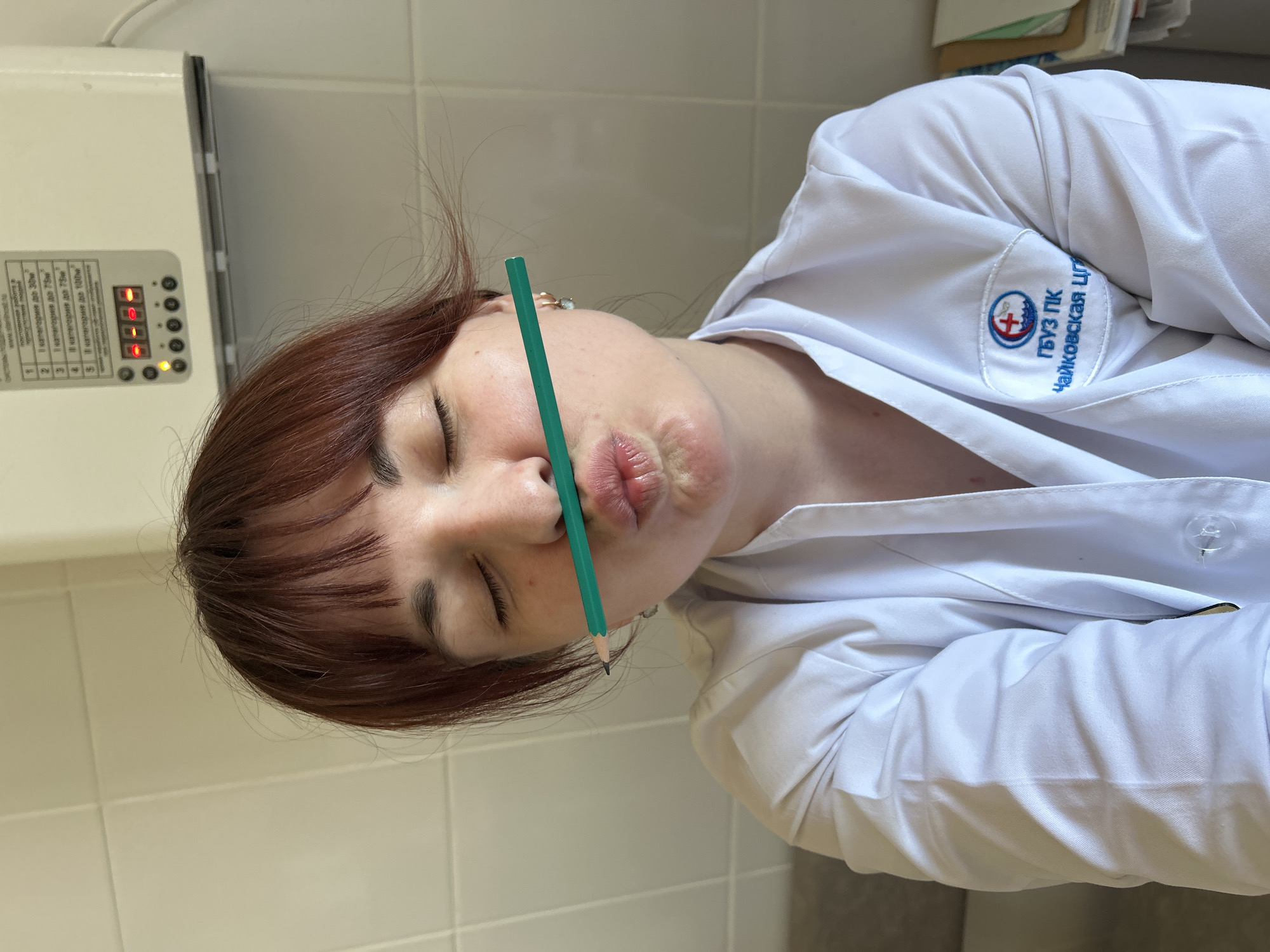 Here is a detailed description of the image:
**Subject:** The image features a woman with auburn, short hair and bangs. She is wearing a white medical coat with a patch on the sleeve that says "ГБУЗ ПК Чайковская цг". She has her eyes closed and is holding a green pencil horizontally between her nose and lips, making a pouting face.
**Environment:** The background appears to be a medical or laboratory setting. A piece of medical equipment with a control panel is visible to the left. The wall behind the woman is made of off-white tiles.
**Objects:** The prominent objects include the green pencil, the woman's medical coat, and the medical equipment.
**Actions/Interactions:** The woman is holding the pencil and making a facial expression.
**Location:** Given the patch on the woman's coat reads "Чайковская цг" ("Chaykovskaya CG"), the location is likely in Chaikovsky, Russia. The Cyrillic letters "ГБУЗ ПК" stand for State Budgetary Healthcare Institution of the Perm Region. Therefore, the photo was most likely taken at the State Budgetary Healthcare Institution of the Perm Region in Chaikovsky, Russia. There is no Фоки mentioned.