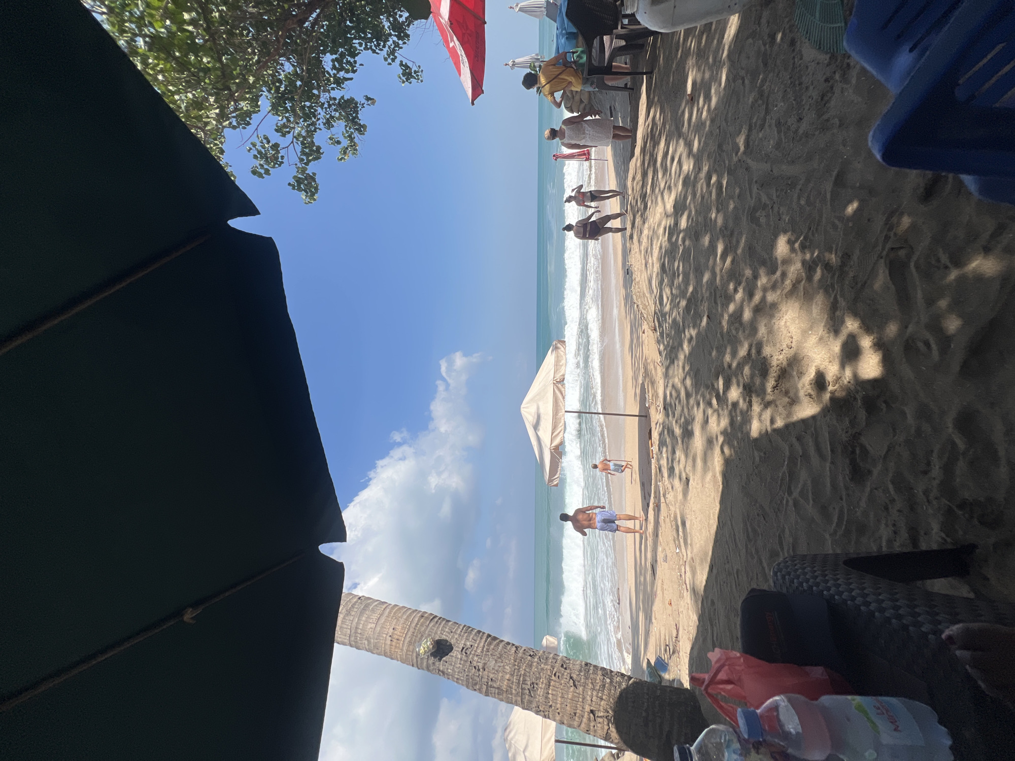 The image captures a sunny day at Kuta Beach, Indonesia, as seen from beneath a dark green beach umbrella. The foreground is dominated by sand and shadows cast by the umbrella and palm trees. To the left, a palm tree trunk stands prominently. In the mid-ground, several people are visible walking on the sandy beach near the water's edge. A man in blue shorts and a child in light shorts are standing near the ocean, which has moderately sized waves. Two women in swimwear and another woman in a white dress are walking further down the beach. To the right, a red umbrella and chairs suggest a seating area. The ocean is a vibrant turquoise, and the sky is a clear blue with scattered white clouds. The scene conveys a relaxed beach atmosphere.