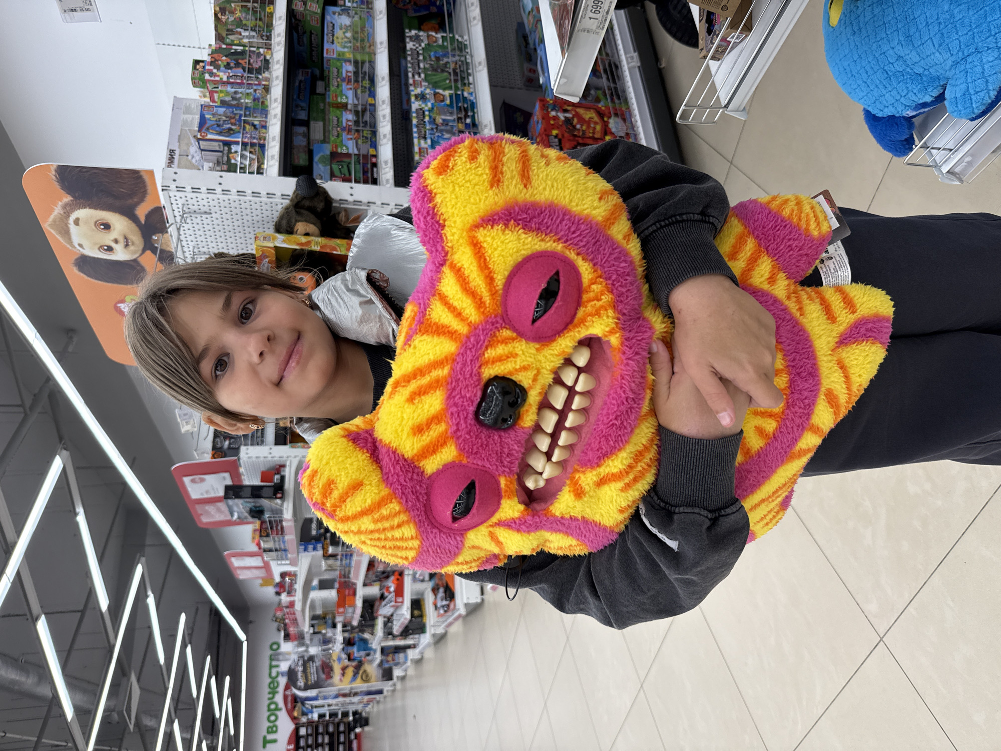 A young person with shoulder-length brown hair smiles at the camera while holding a large, brightly colored stuffed toy. The toy is yellow and pink with a fierce expression, featuring large black eyes, sharp white teeth, and a pink mouth. The person is wearing a dark gray hooded sweatshirt and dark pants. They are standing in what appears to be a store aisle with shelves stocked with toys and other merchandise in the background. The store lighting is overhead and fluorescent. The word "Творчество" (meaning "Creativity" in Russian) is visible on a green sign in the background, suggesting the store is a toy or craft store. The floor is tiled with a beige, light-colored pattern. The overall environment is a retail setting, likely in Yaroslavl, Russia, given the Cyrillic signage.