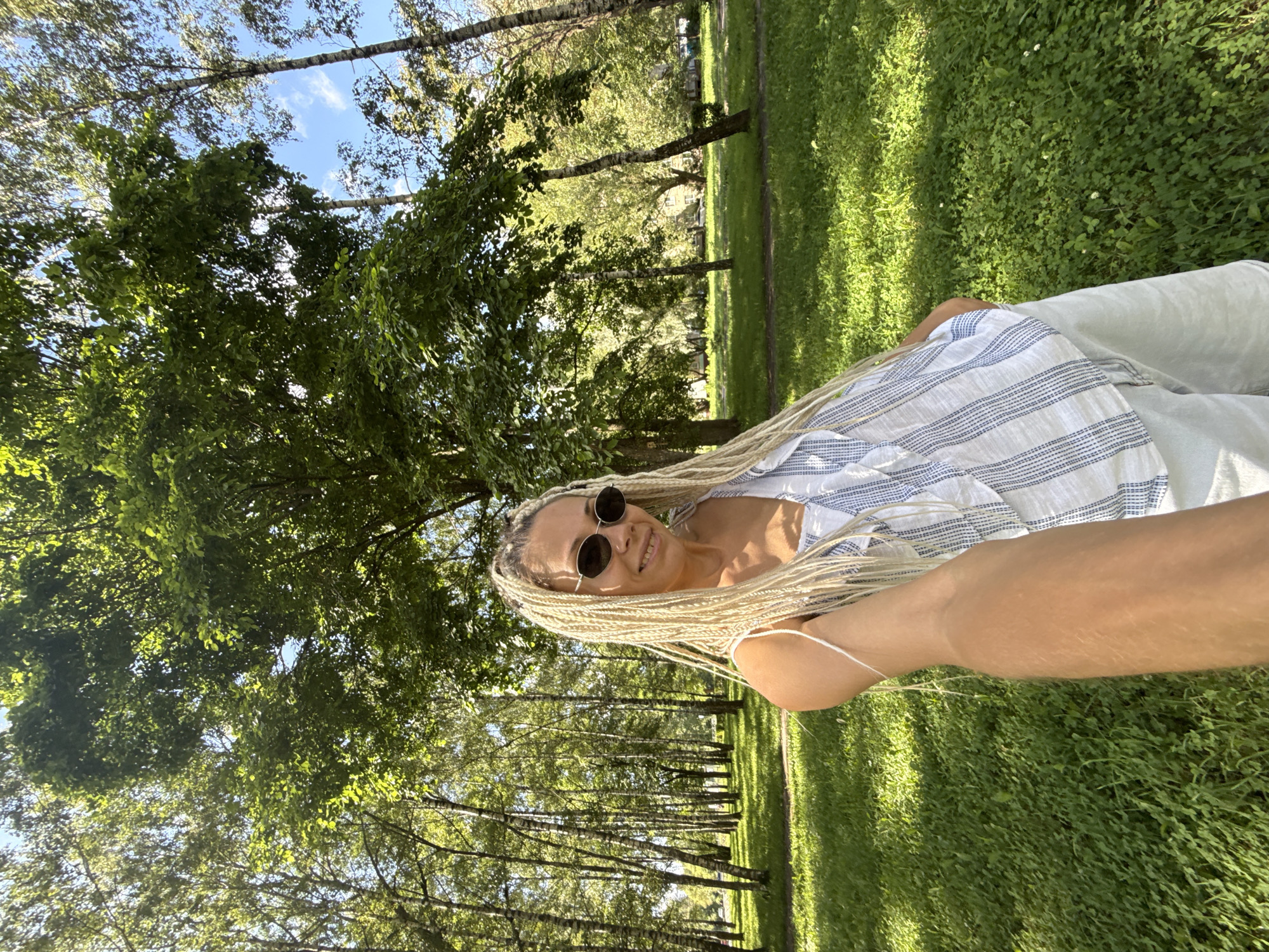 A woman with long blonde braids is taking a selfie in a park in Rusanovo, Russia. She is wearing sunglasses, a white and blue striped top, and white shorts. The park is filled with trees and green grass, and the sun is shining.