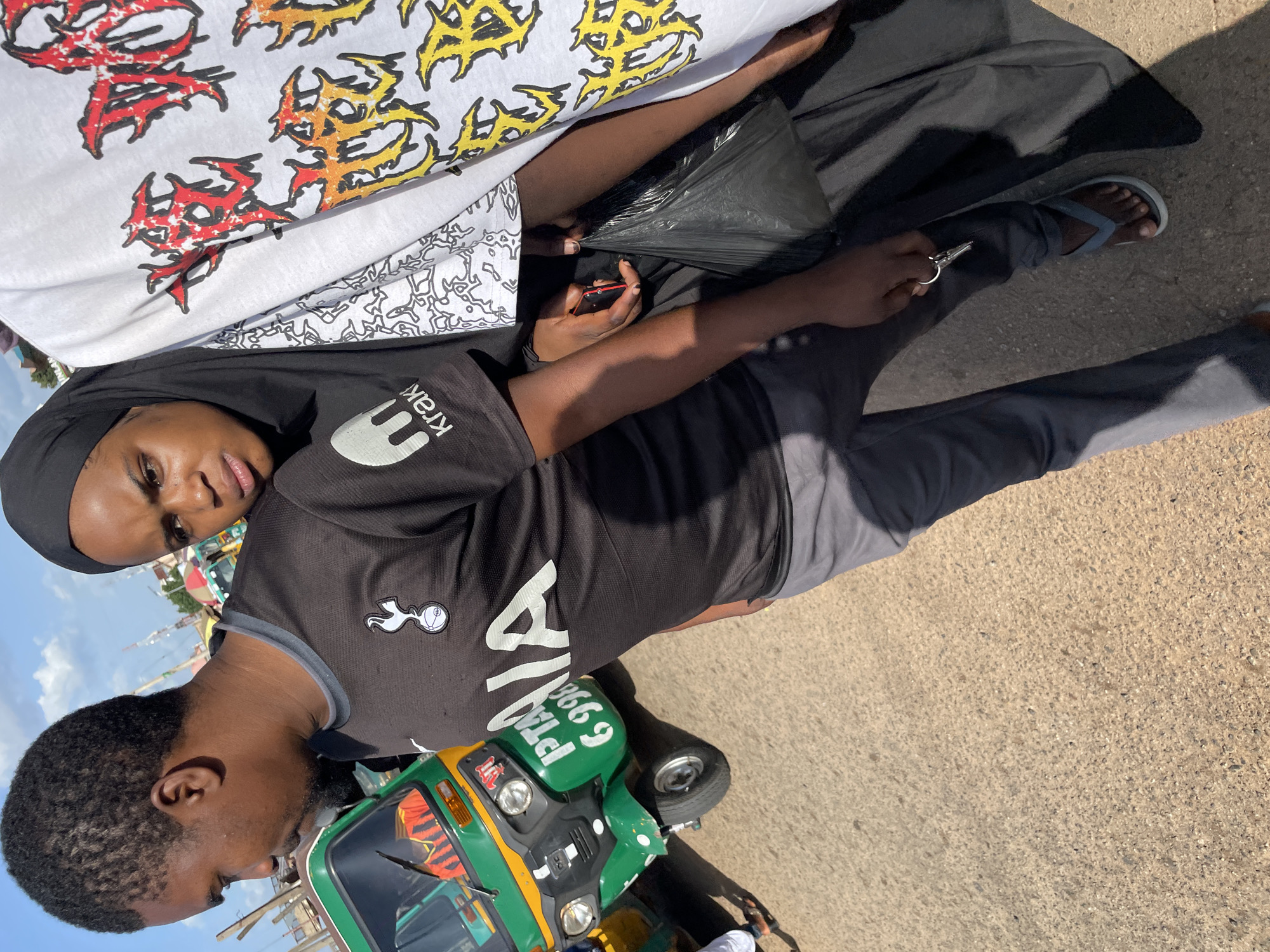 A man and a woman are standing next to a green auto-rickshaw in what appears to be a market or busy street. The man is wearing a black t-shirt with a white logo and grey pants, and the woman is wearing a black headscarf and a black dress. The auto-rickshaw has "PTA 6998" written on its side in white lettering. There are other vehicles and structures in the background, suggesting a bustling urban environment. The scene is well-lit, indicating it is daytime. The image captures a candid moment of these individuals in their surroundings.
