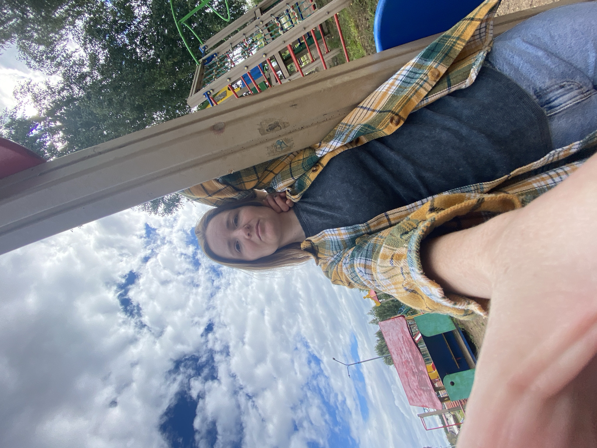 A woman with blonde hair is taking a selfie on a playground. She is wearing a dark grey ribbed top, a yellow, green, and white plaid shirt, and blue jeans. She is leaning against a wooden pole. In the background, there is a playground structure with red and green elements, a blue plastic seat, and trees. The sky is cloudy with patches of blue. The image was taken in Chaikovsky, Russia, likely on a playground.