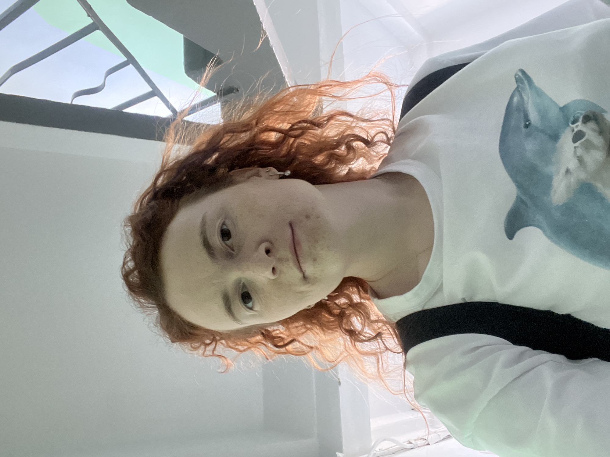 A woman with curly red hair, freckled skin, and fair complexion, is wearing a white t-shirt with a graphic of a blue dolphin. The photo is taken from a low angle, looking up towards the woman. In the background, there is a structural metal frame, possibly part of a building or skylight. The location is potentially within the city of Chaikovsky, Russia, but this is not definitively clear from the image. The environment is bright and appears to be indoors.