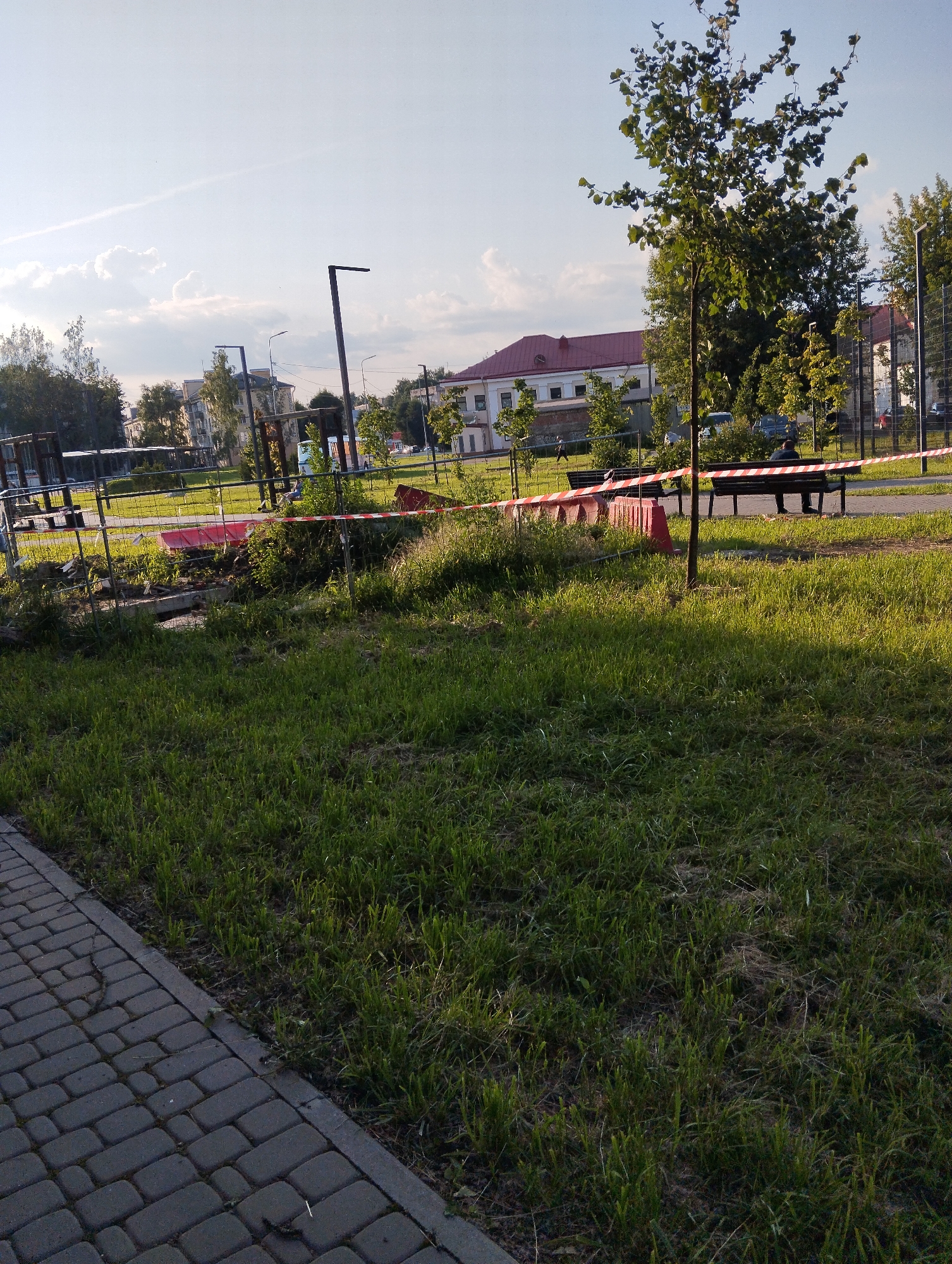 The image depicts a park area in Kingisepp, Russia, with a paved walkway in the foreground. A section of the park is cordoned off with red and white striped tape and orange traffic cones, indicating a construction or maintenance zone. Within the restricted area, there is disturbed earth, some debris, and what appears to be a red rectangular object. Beyond the fenced-off area, there are park benches, one occupied by a person sitting, and a young tree. In the background, buildings with red roofs are visible, as well as other trees and a fence. The sky is partly cloudy, with the sun casting shadows.